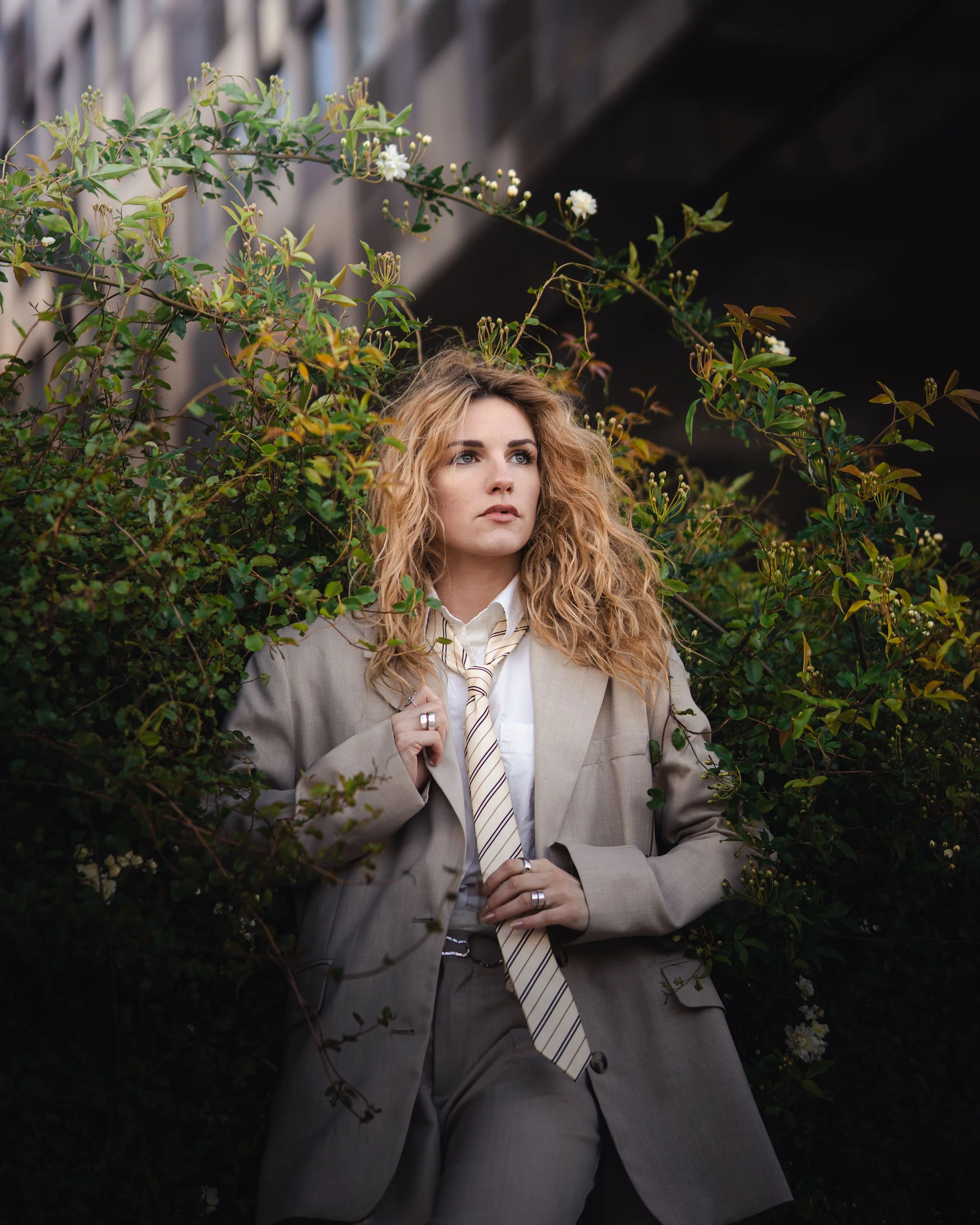 Une femme aux cheveux bouclés, habillée d'un costume beige avec une chemise blanche et une cravate rayée, posant près de buissons verts et de fleurs blanches.