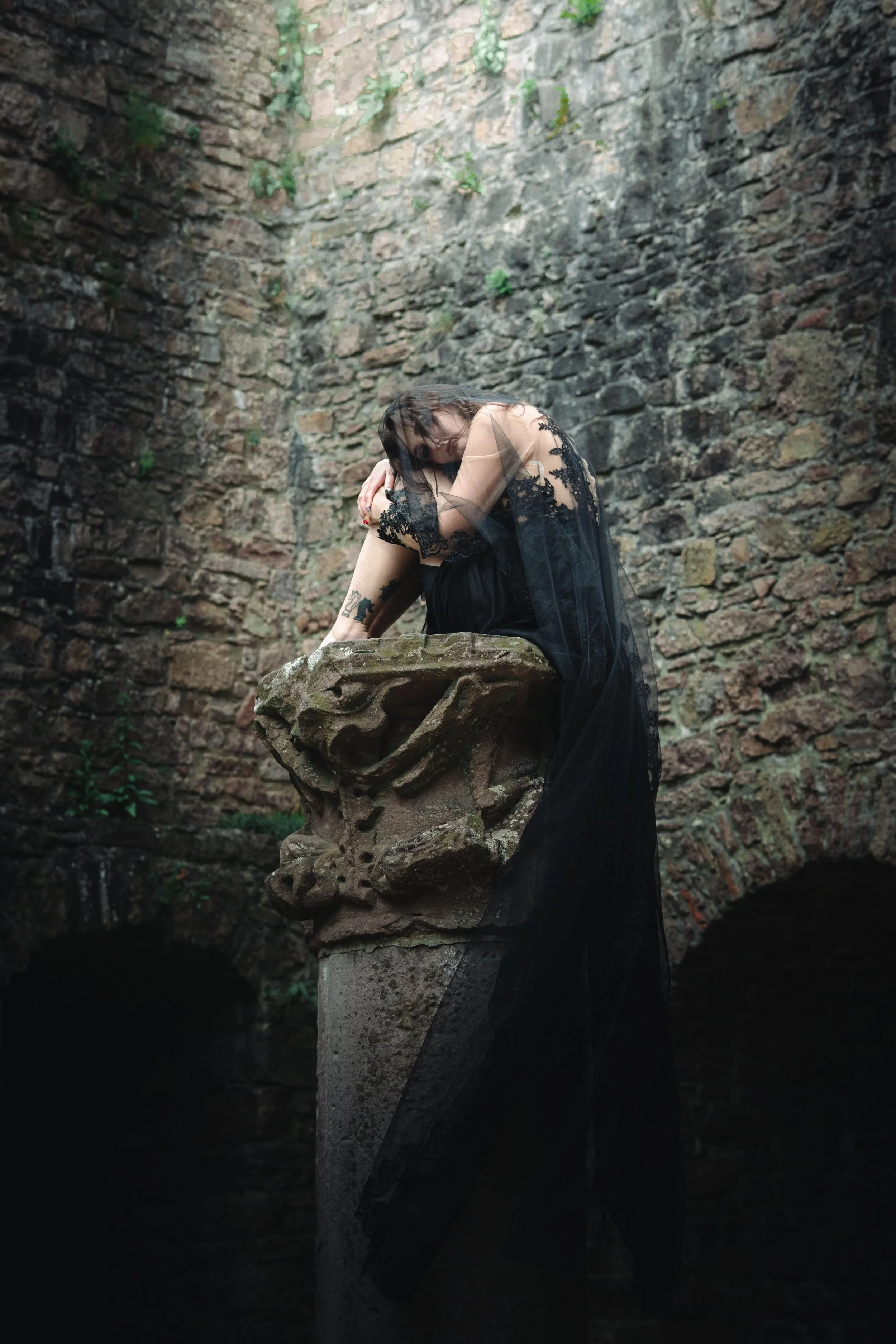 Portrait d'une femme portant une robe noire en dentelle et un voile noir posé sur ses épaules, positionnée sur une sculpture en pierre dans un environnement rocheux avec un mur de pierres derrière, au château de Hohenbaden.