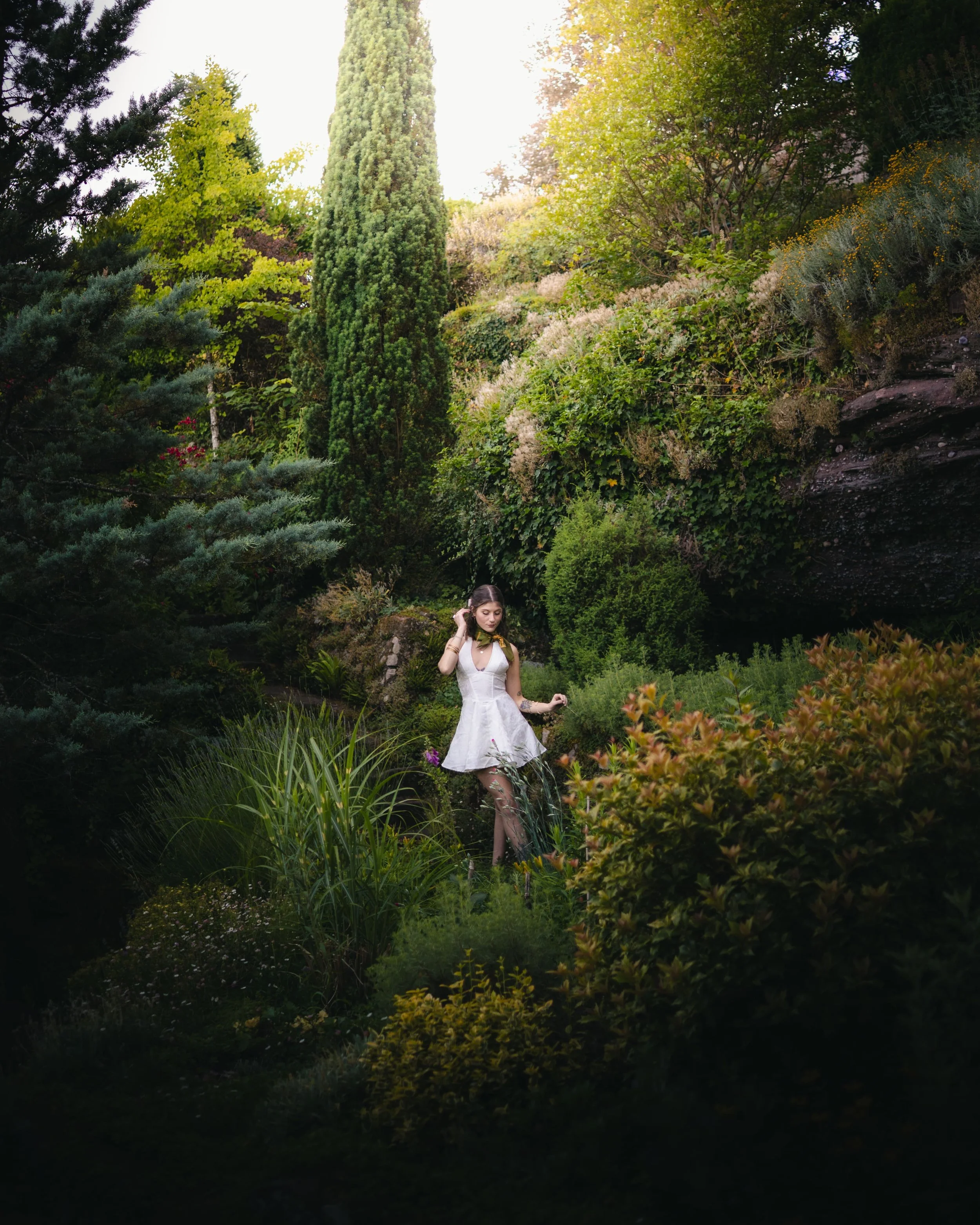 Portrait d'une femme en robe blanche dans un jardin dans les Vosges avec diverses plantes, arbres, et feuillage dense.