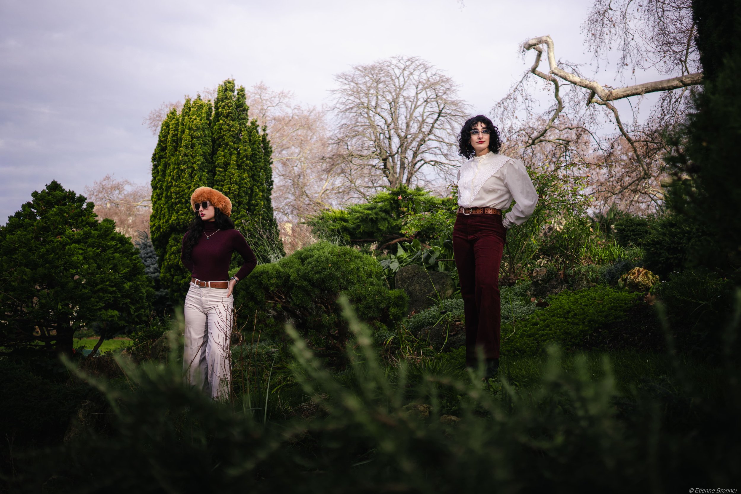 Deux femmes posent dans un jardin avec des arbres et des buissons, portant des vêtements vintage.