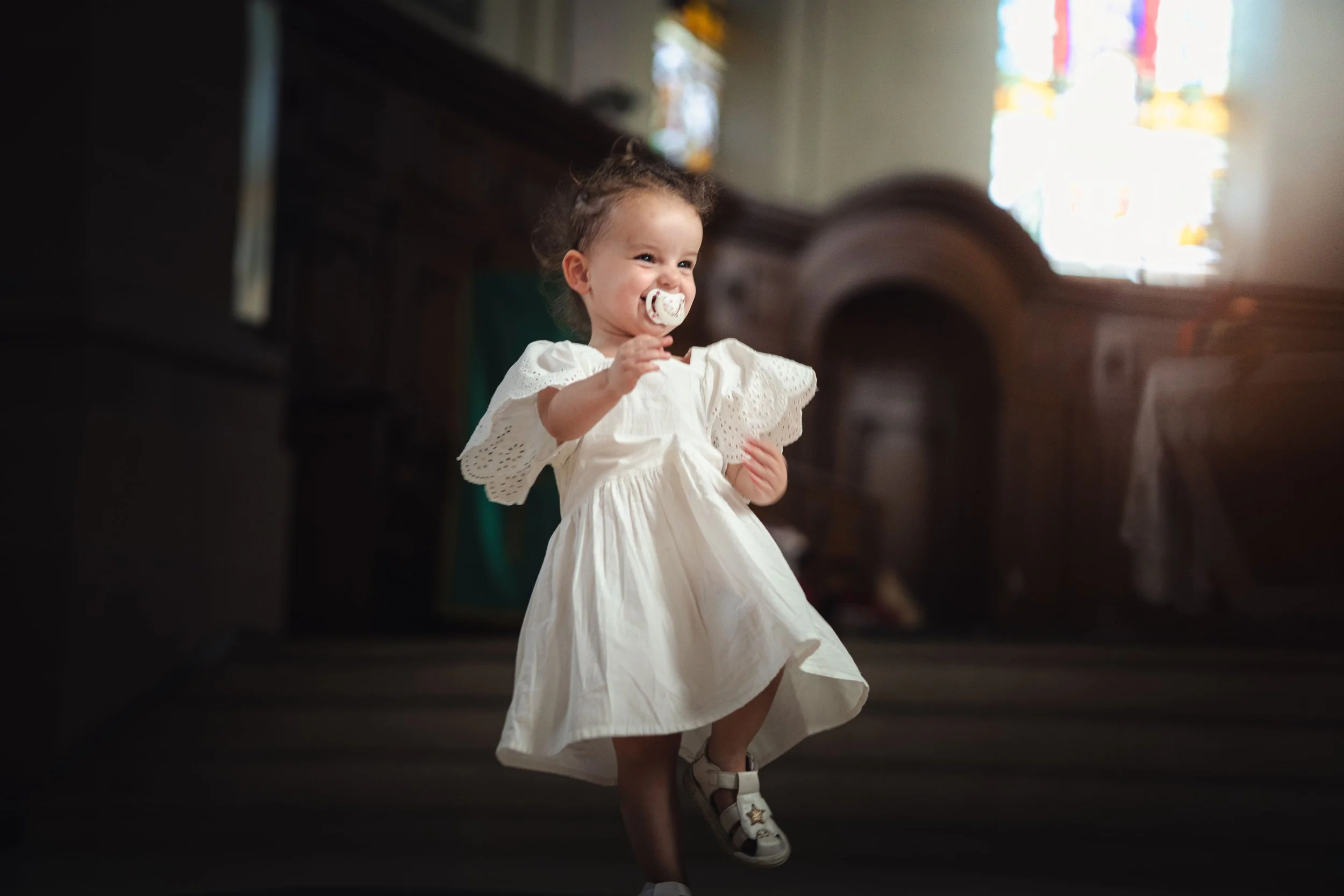 Portrait d'une petite fille souriante portant une robe blanche avec des dentelles, marchant dans une église ou une salle de cérémonie, avec des vitraux colorés en arrière-plan.