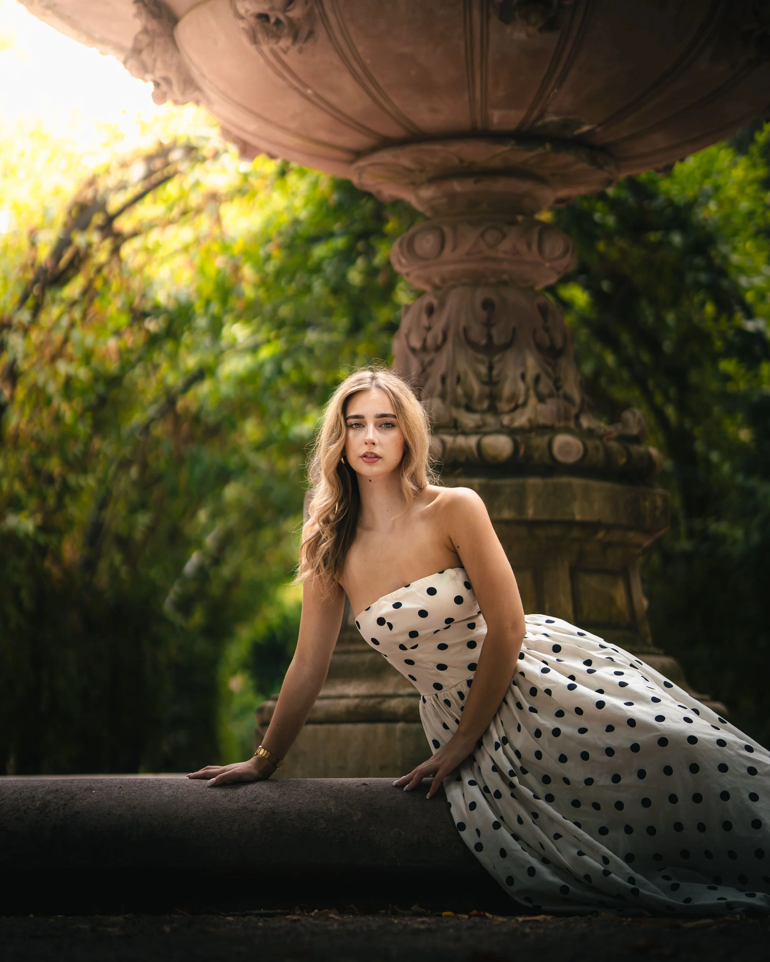Portrait d'une femme en robe blanche à pois noirs est agenouillée près d'une fontaine décorative en pierre dans un jardin vert, au parc de l'orangerie à Strasbourg.