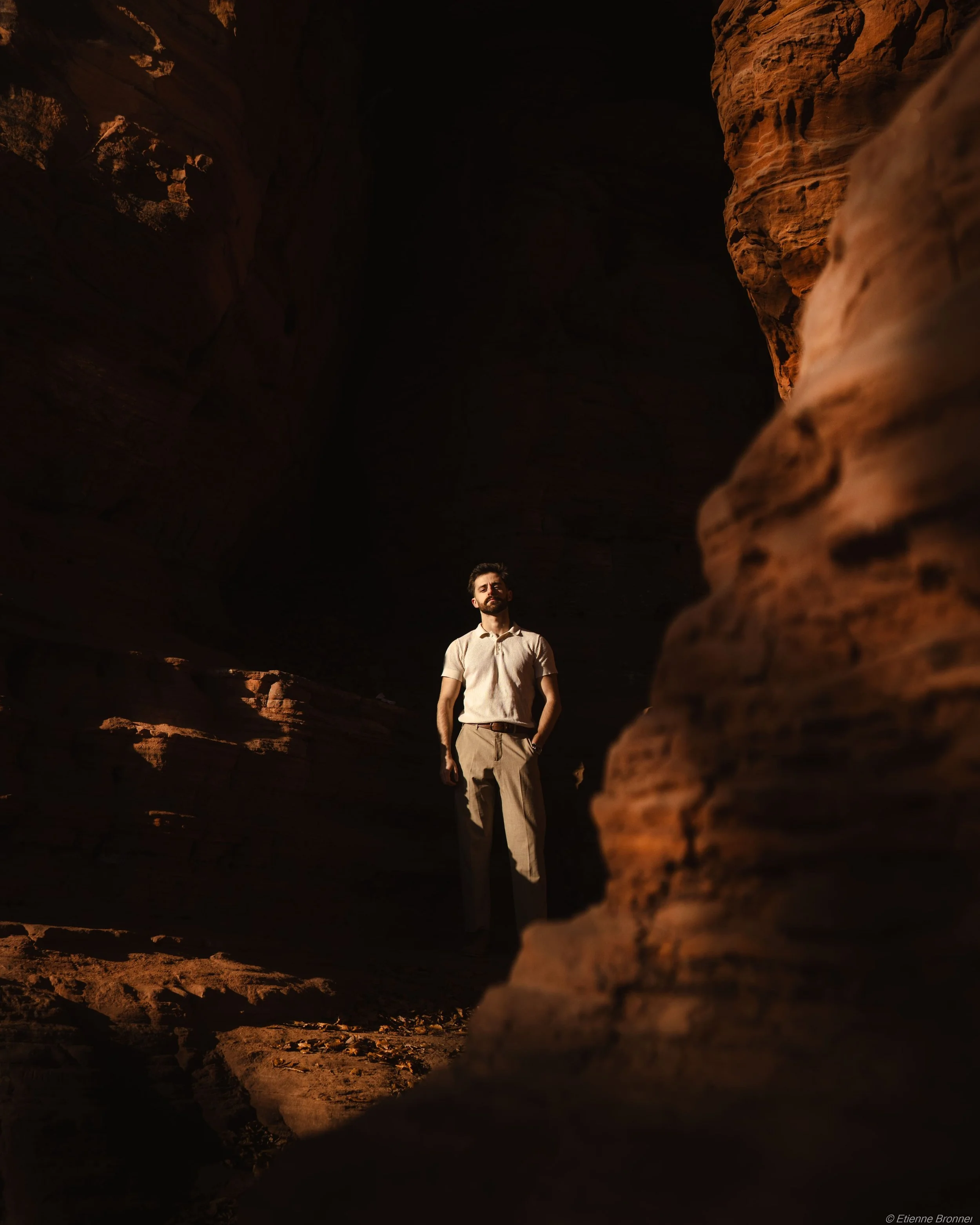 Homme debout dans une grotte sombre avec des parois rocheuses orange, éclairé par une lumière naturelle
