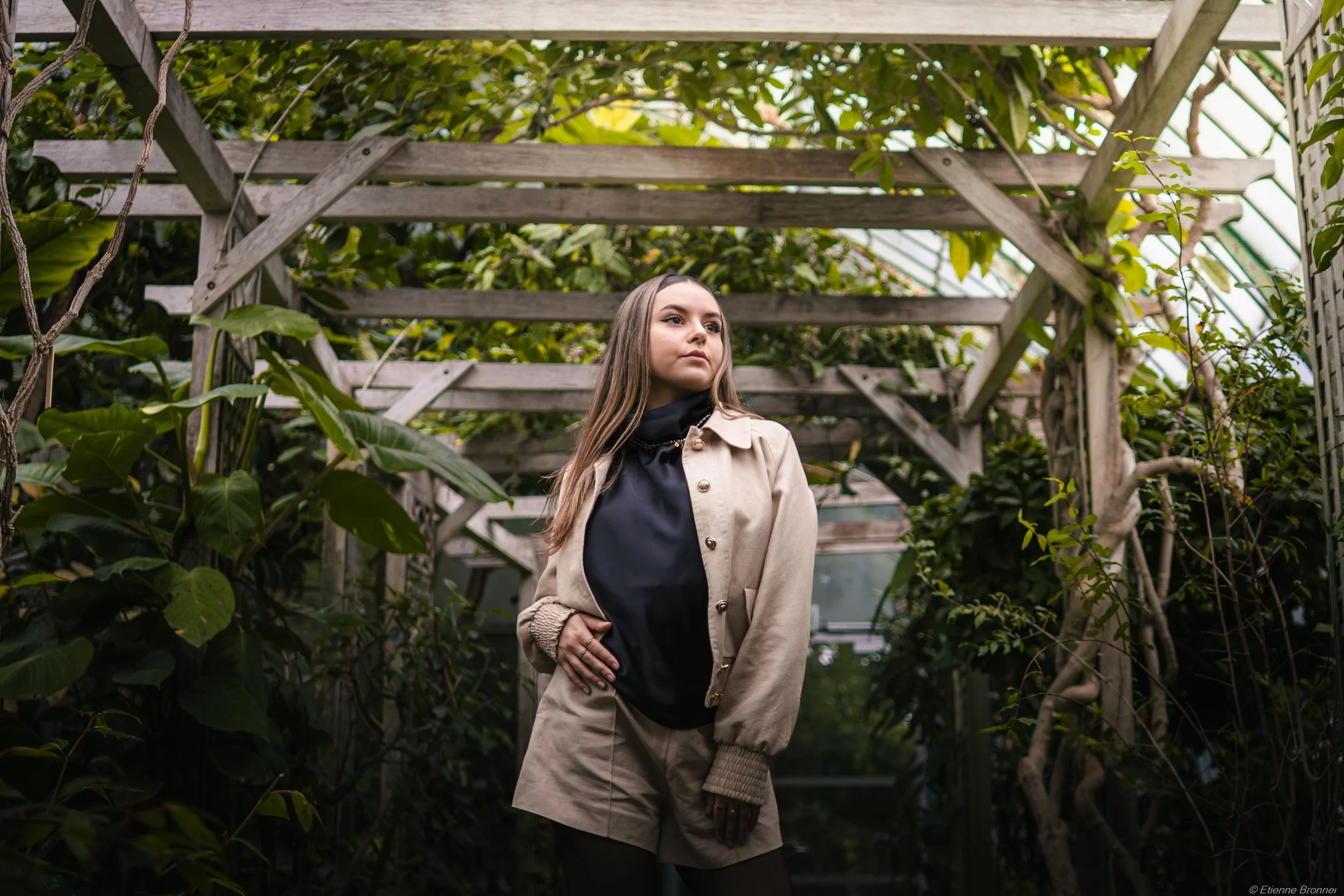 Jeune femme dans une serre végétale, les serres d'Auteuil, portant un blazer beige, une robe noire, et des gants, entourée de plantes vertes et de structures en bois.
