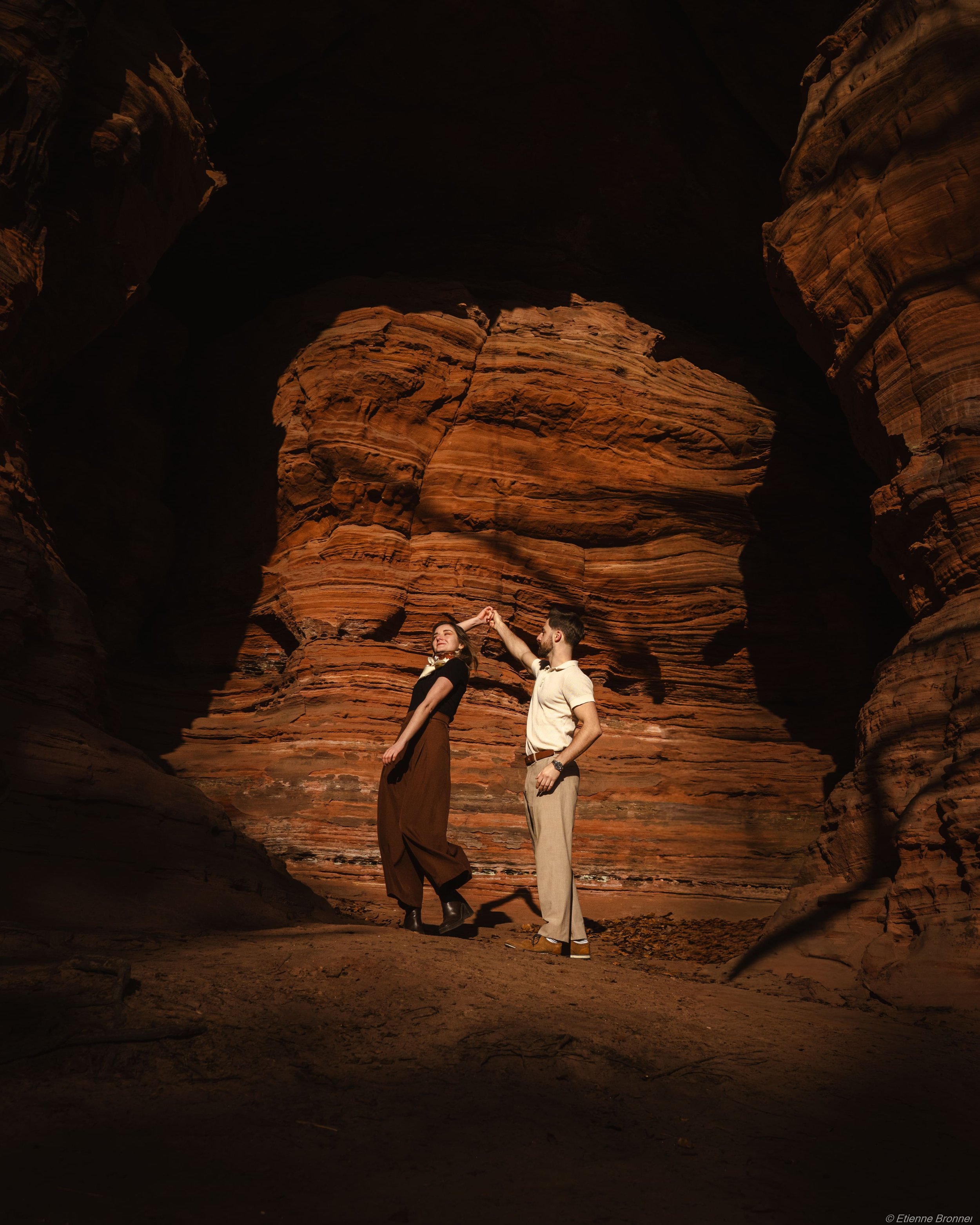 Deux personnes dans une formation rocheuse rouge, une femme et un homme, se tiennent et se touchent la main. La scène est dans une sorte de canyon ou grotte ensoleillée.