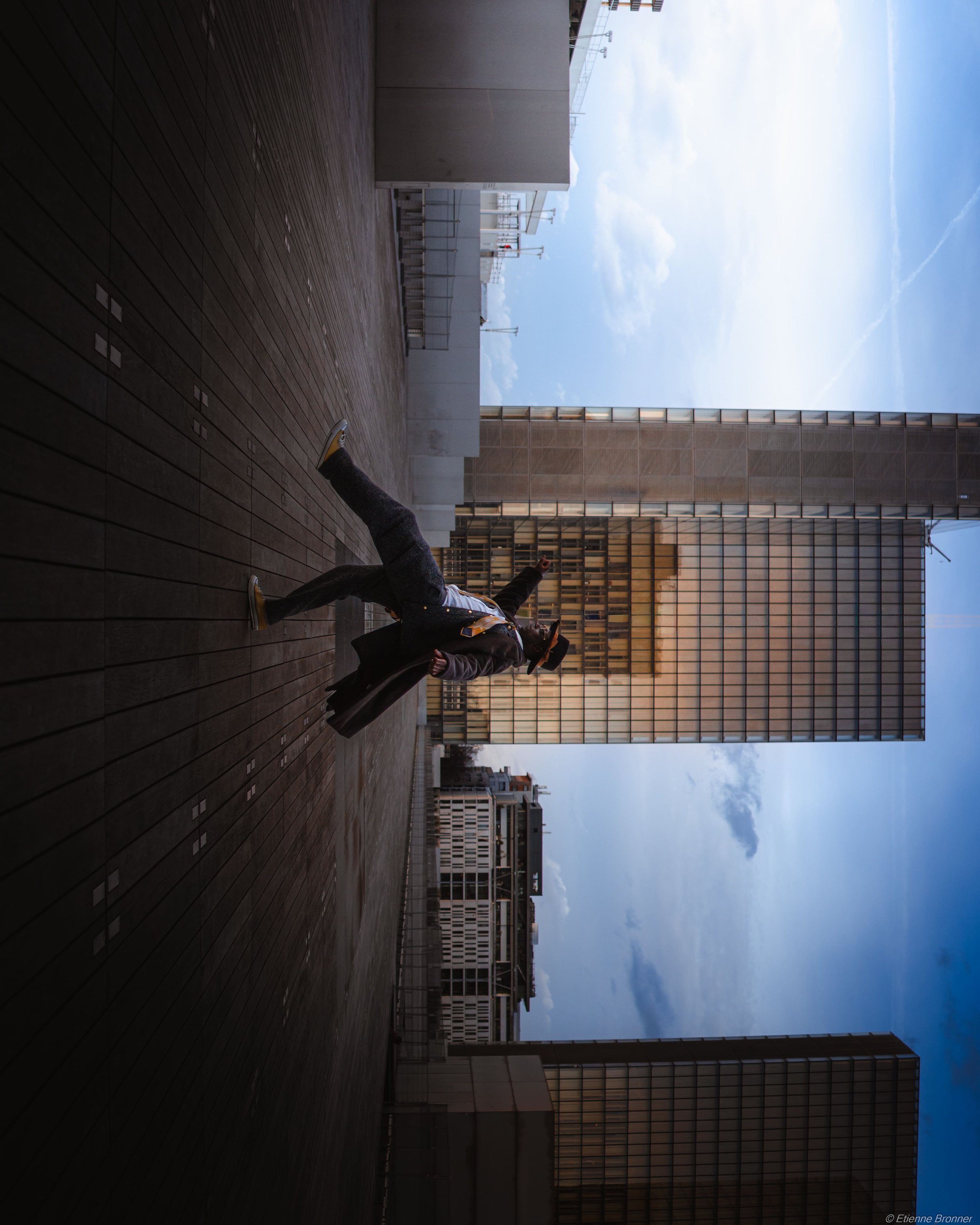 Une personne danse ou fait une pose dynamique sur un toit ou une terrasse en milieu urbain, avec plusieurs bâtiments modernes en arrière-plan et un ciel en partie nuageux.