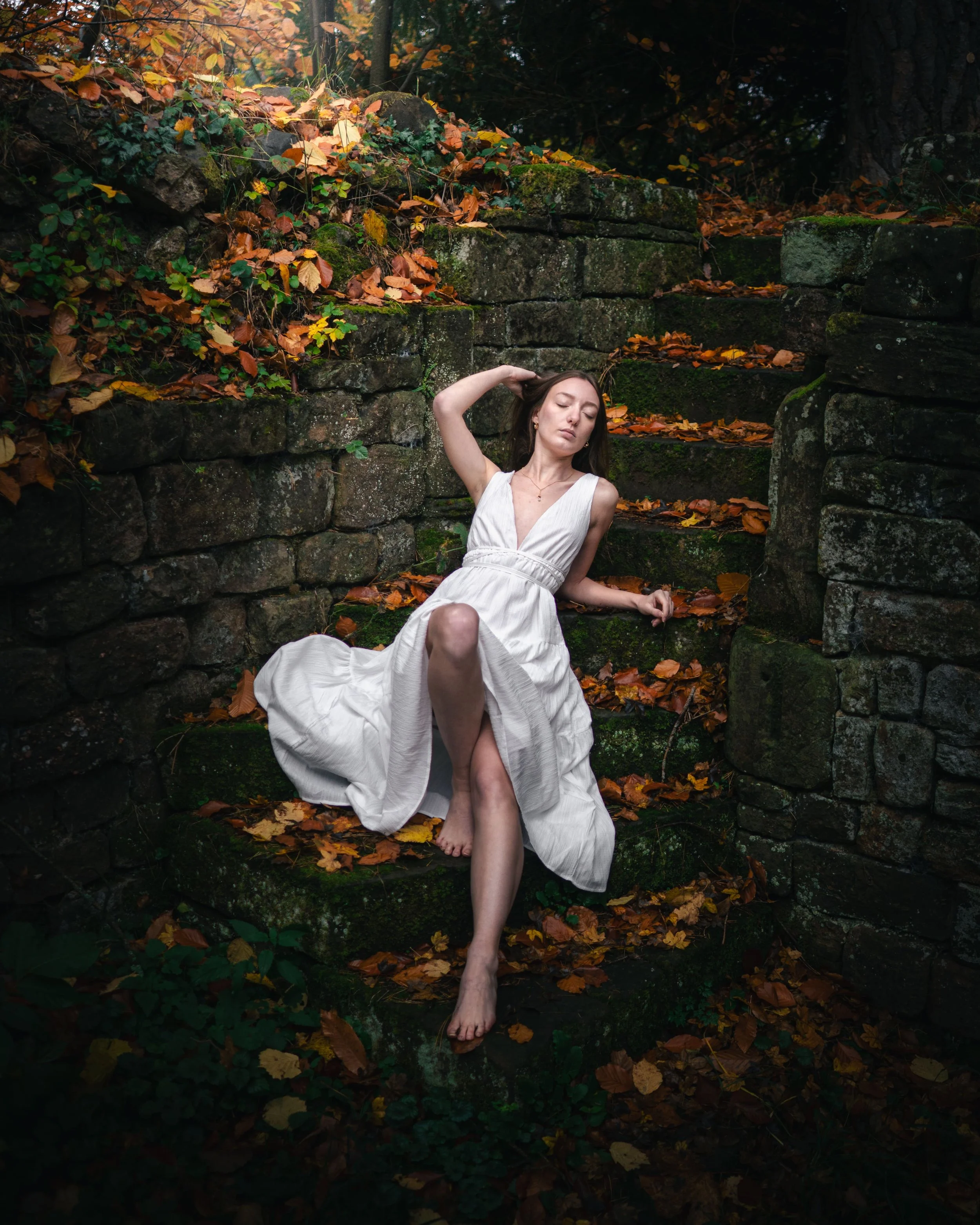 Portrait d'une femme en robe blanche assise sur des escaliers en pierre couverts de feuilles mortes automnales dans une forêt, au domaine du Windeck à Ottrott.