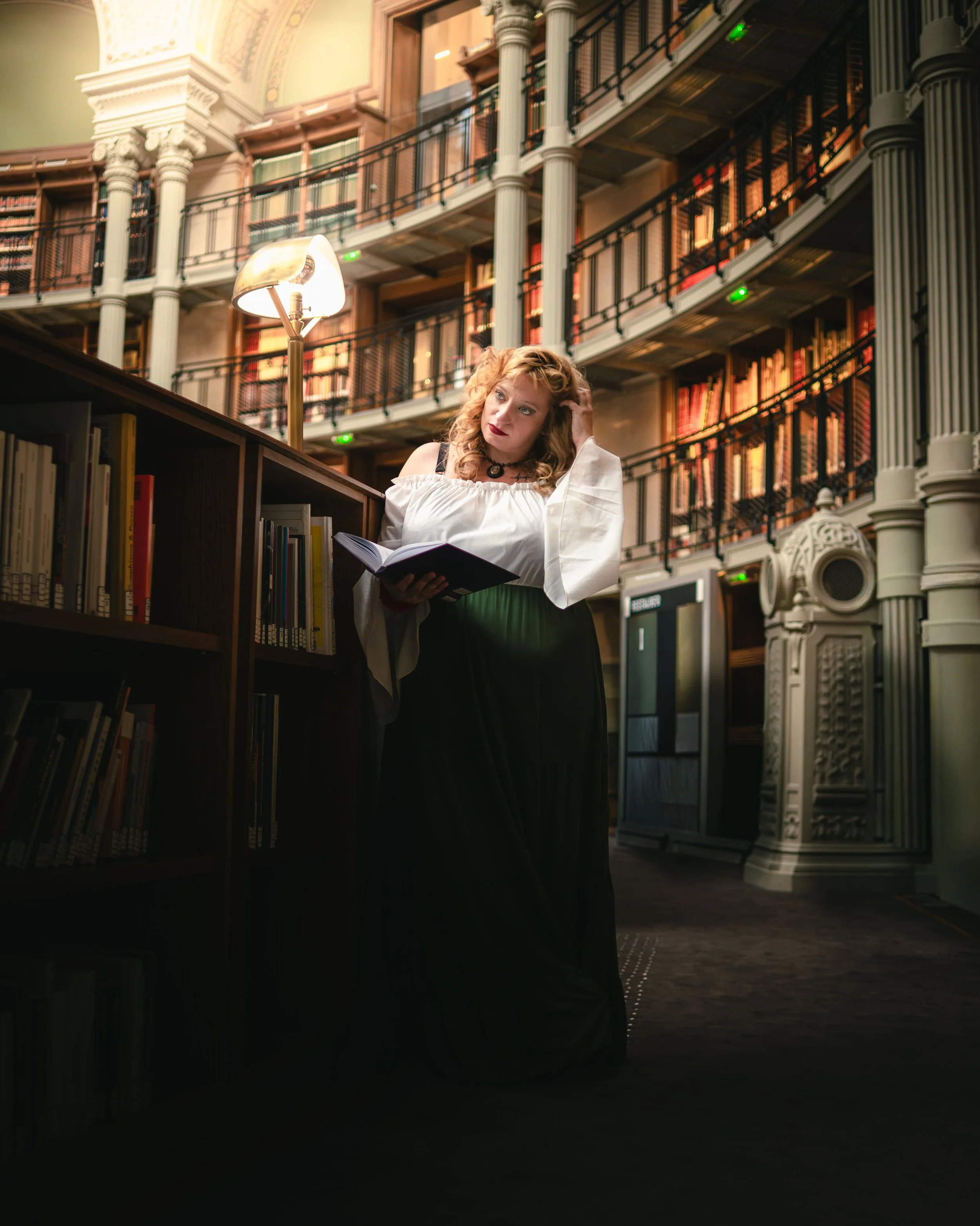 Portrait d'une femme avec des cheveux bouclés roux, portant une robe blanche et un collier noir, consulte un livre dans une bibliothèque ancienne avec plusieurs étages, des lampes et des meubles en bois, à la BNF de Paris.