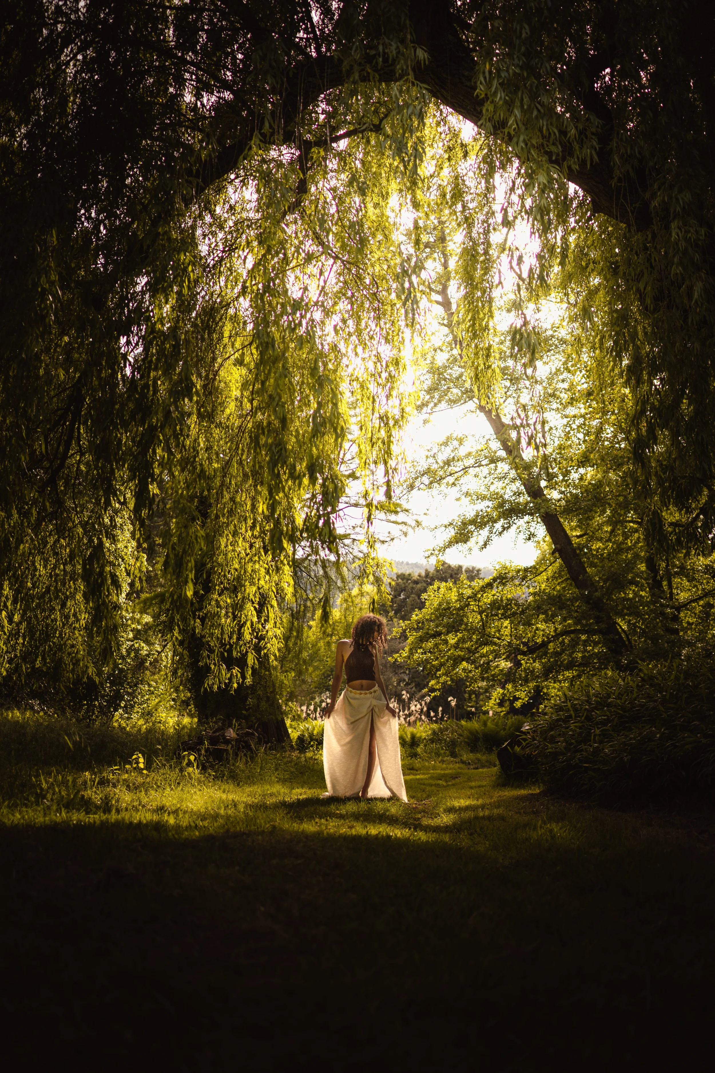Portrait d'une femme debout dans un jardin au domaine du Windeck en plein air entouré d'arbres, sous la lumière du coucher de soleil. Elle porte une longue jupe claire et un haut noir.