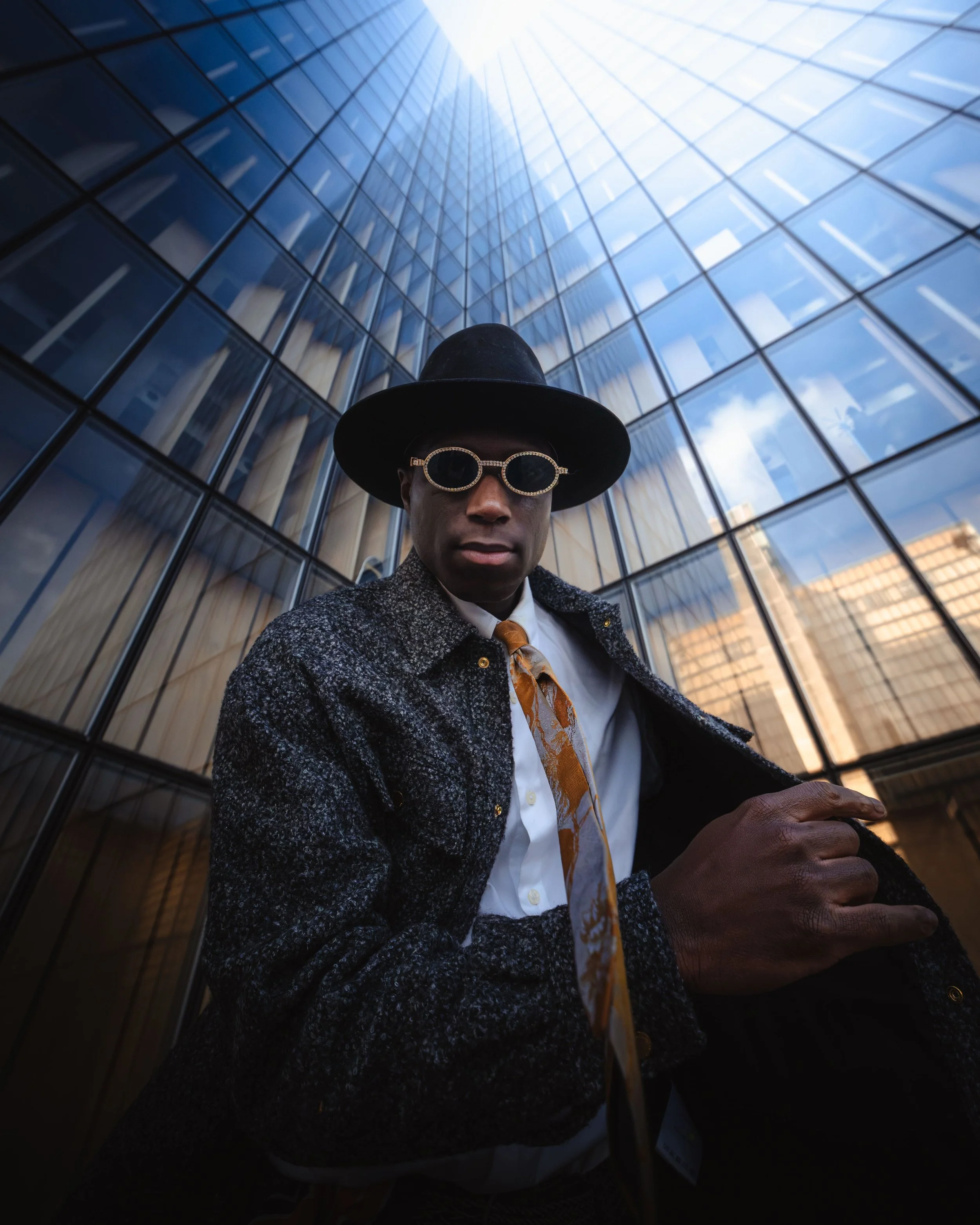 Portrait d'un homme portant un chapeau noir, des lunettes ornées de pierres et un manteau en tweed se tient devant la BNF de Paris, le ciel en arrière-plan.