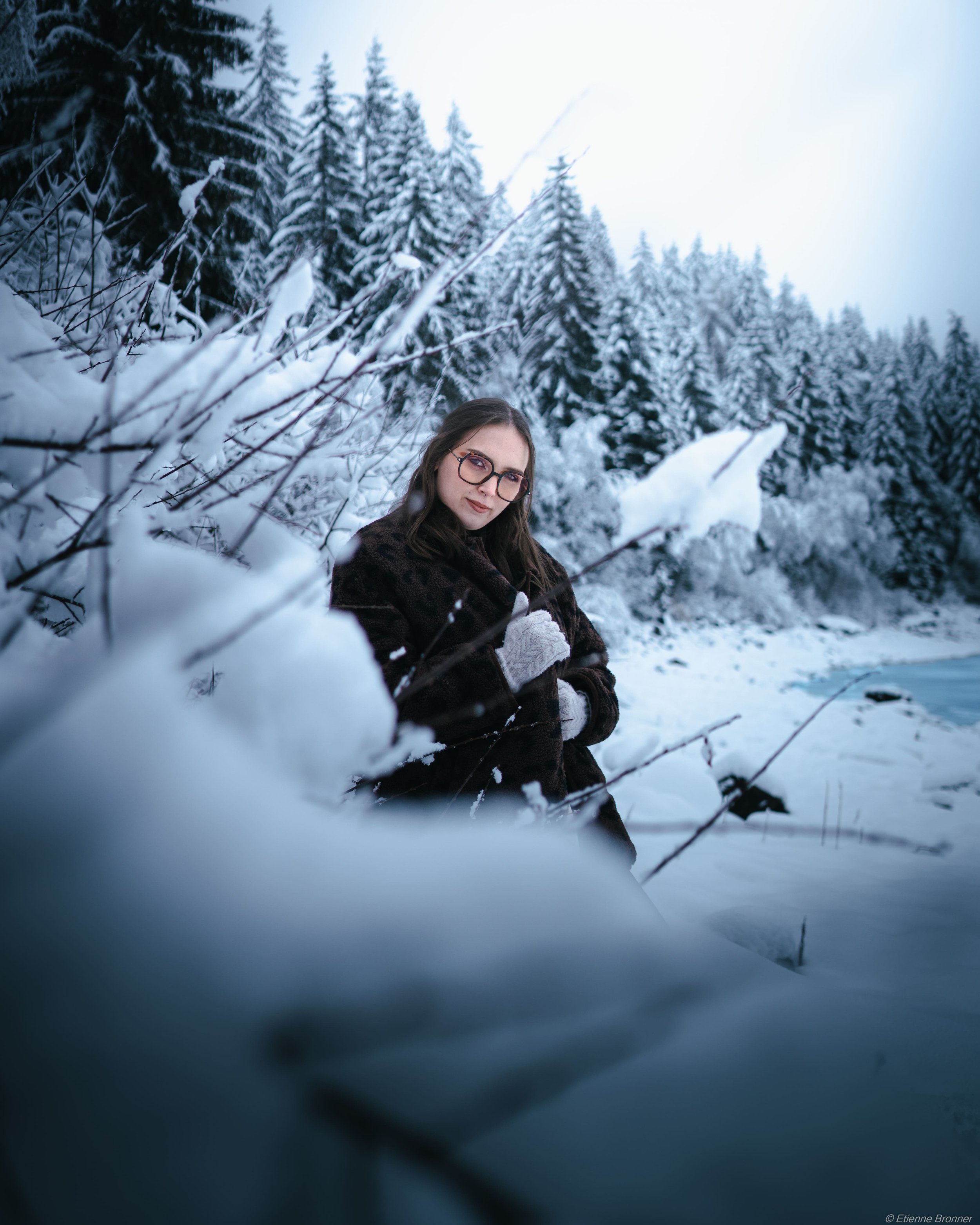 Portrait d'une femme portant des lunettes, vêtue d'une veste brune, se tient dans un paysage enneigé entouré de sapins recouverts de neige, avec un ciel clair en arrière-plan, au lac blanc dans les Vosges.