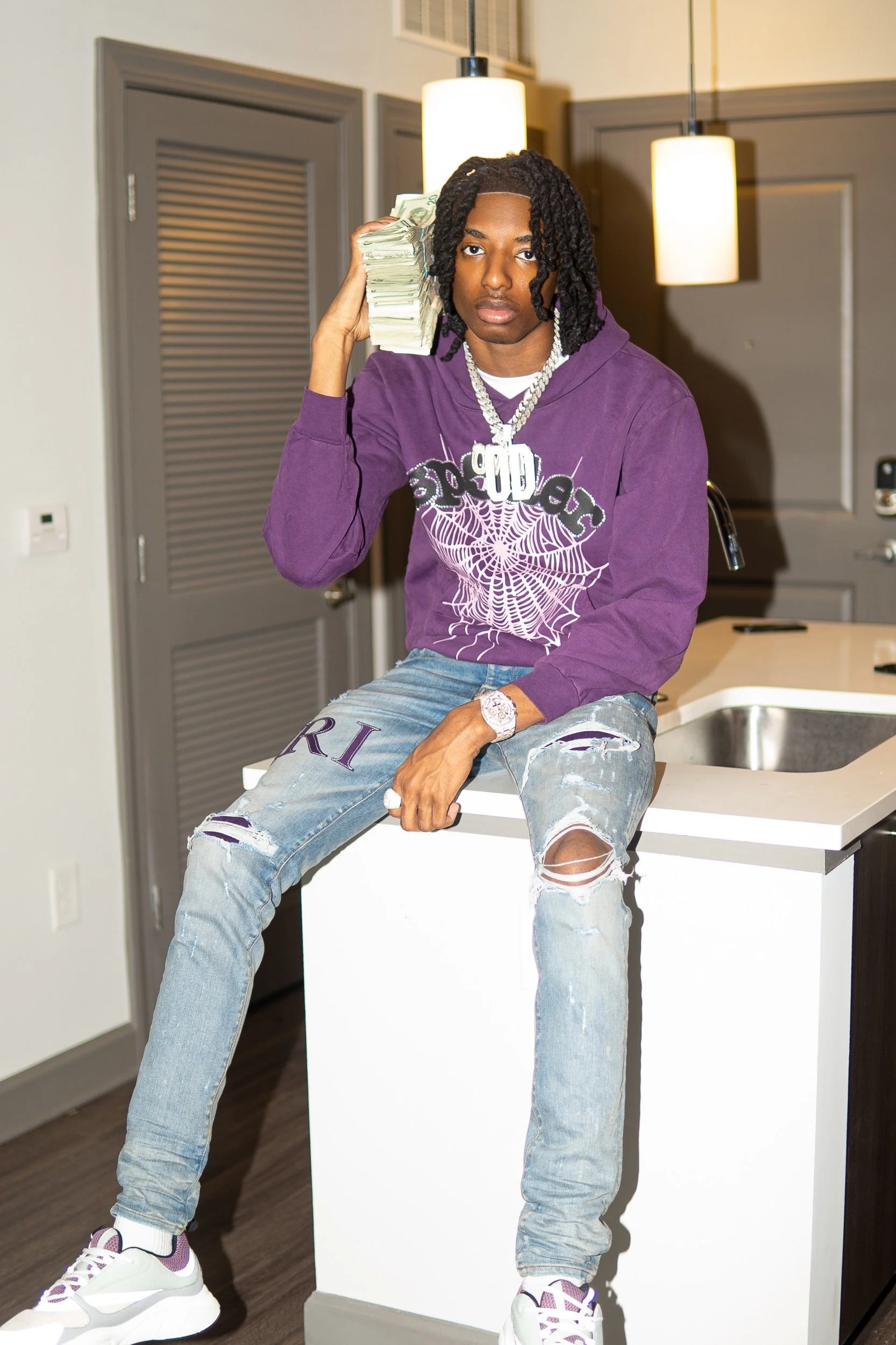 Young man with dreadlocks wearing a purple hoodie, ripped jeans, and white sneakers, sitting on a kitchen counter, holding a large stack of money on his shoulder.