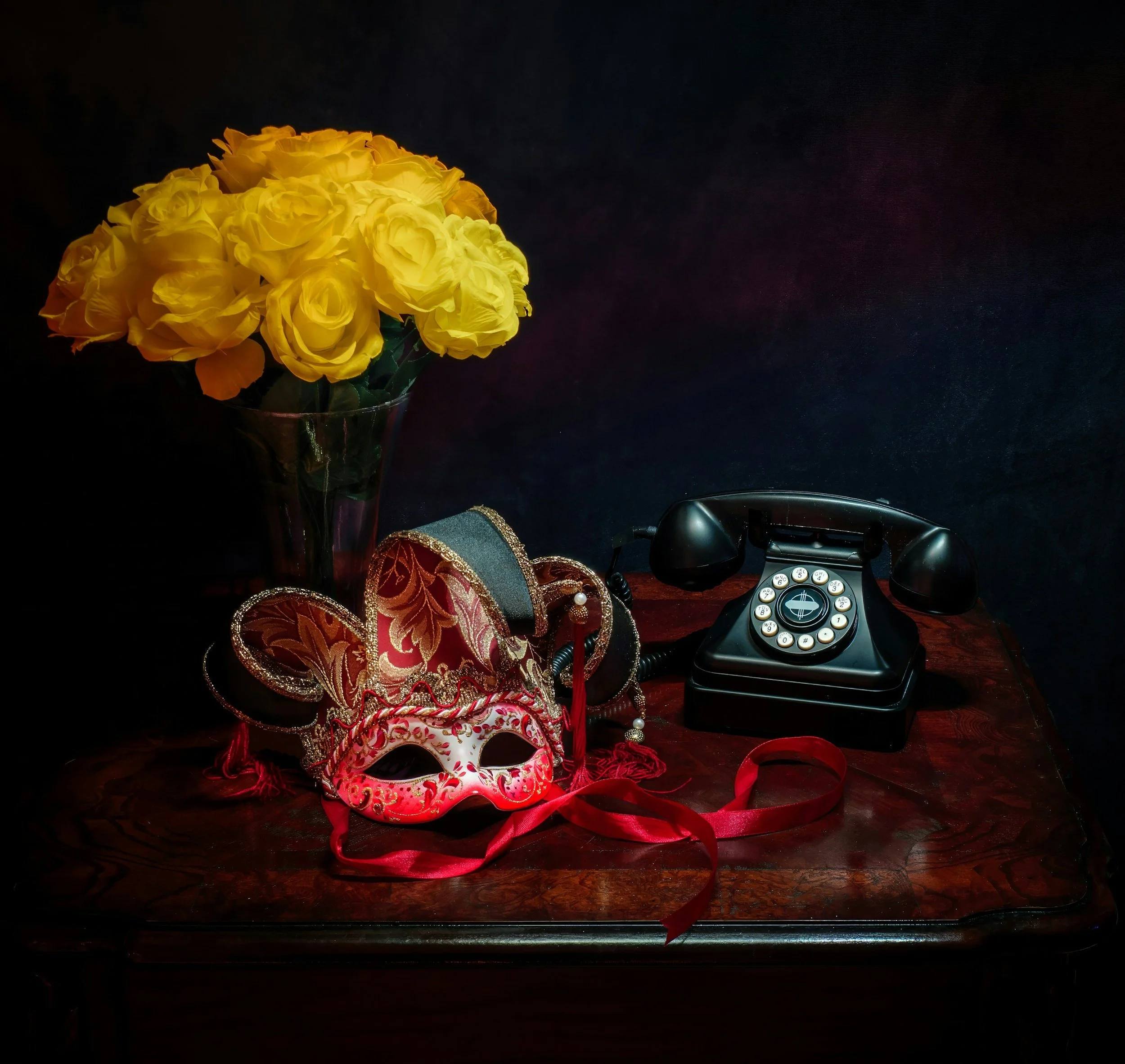 A dark wooden table with a large bouquet of yellow roses in a glass vase, a decorative Venetian mask with red, gold, and teal details, and a vintage black rotary dial telephone.