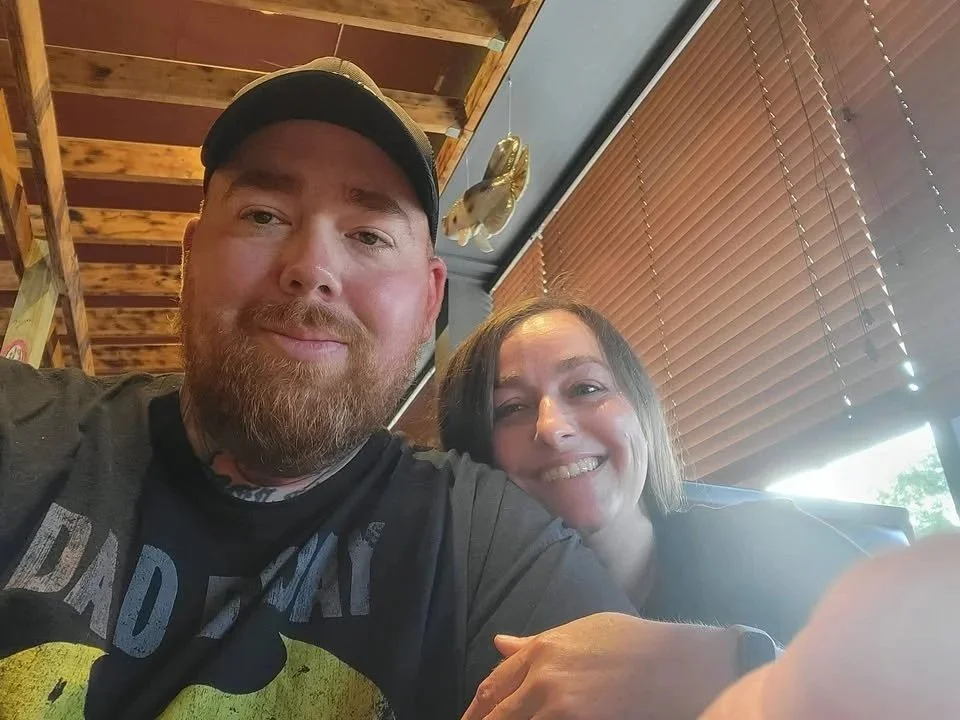 A man wearing a black cap and a black T-shirt sitting in a restaurant with a smiling woman, wooden blinds, and a wooden ceiling in the background.