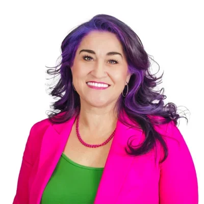 Woman with purple and black hair, wearing a bright pink blazer and green top, smiling.