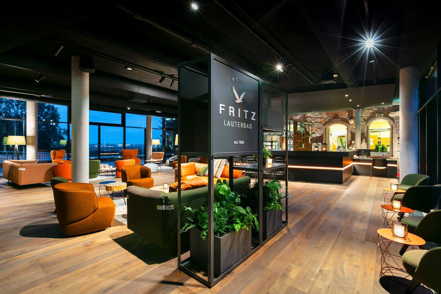 Innenansicht eines modernen Hotel-Lounges mit bequemen bunten Sesseln, großen Fenstern, Pflanzen und einer Rezeption im Hintergrund.