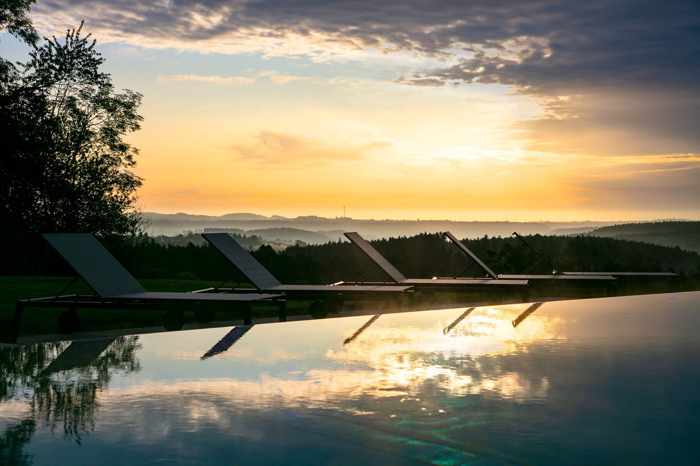 Sonnenuntergang über einer Landschaft mit Bäumen, Blick auf einen Infinity-Pool mit Sonnenliegen und reflektiertem Himmel.