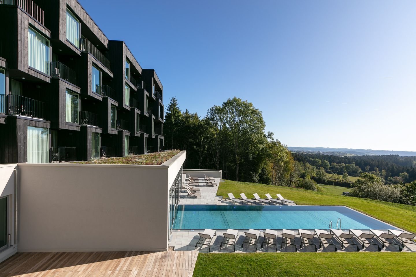Modernes Hotel mit Außenpool, Liegestühlen und Blick auf eine grüne Landschaft unter klarem Himmel.