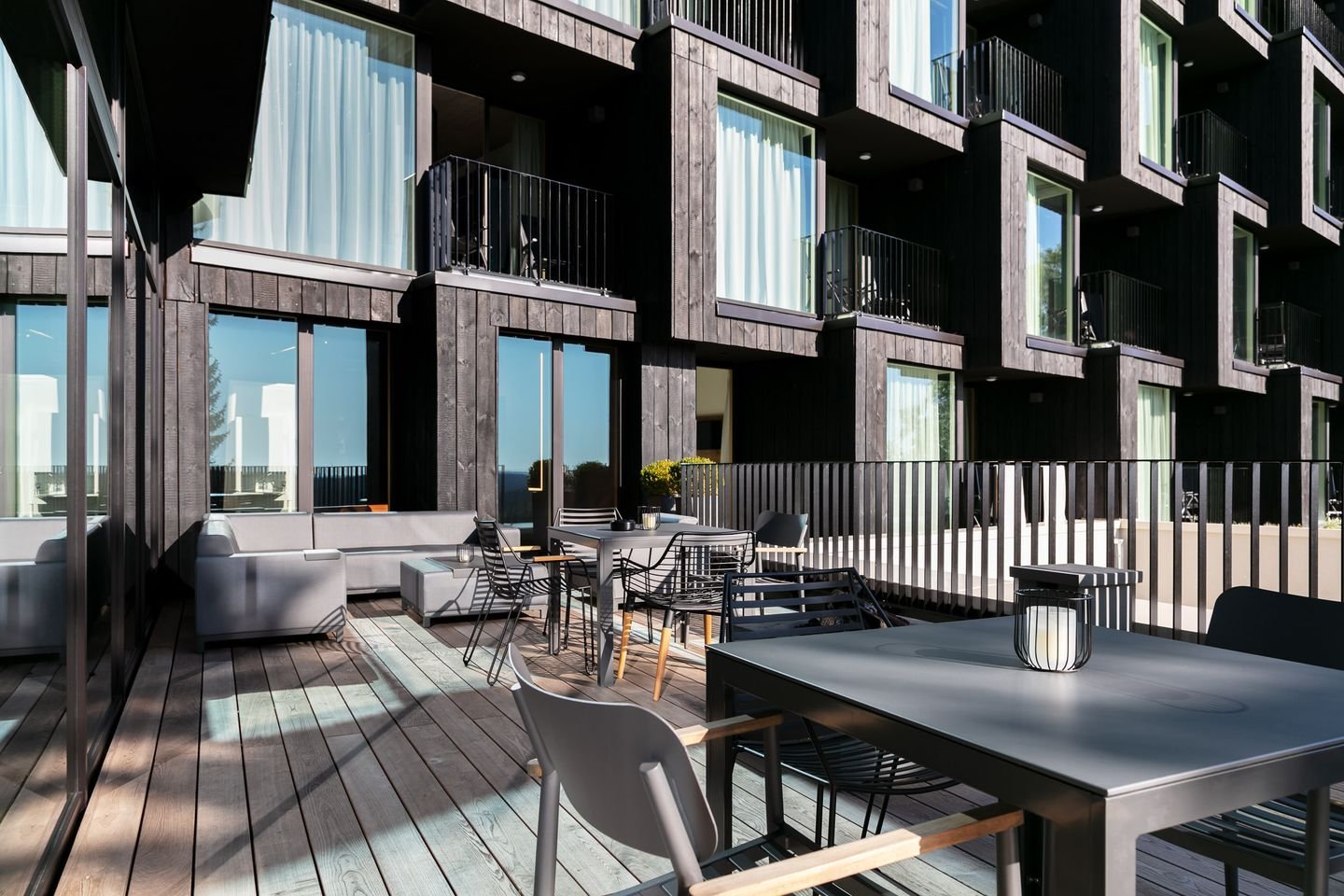 Balkon eines modernen Hauses mit schwarzen Holzverkleidungen, mehreren Tischen und Stühlen, Sonnenschirmen, dekorativen Laternen und Aussicht auf einen blauen Himmel