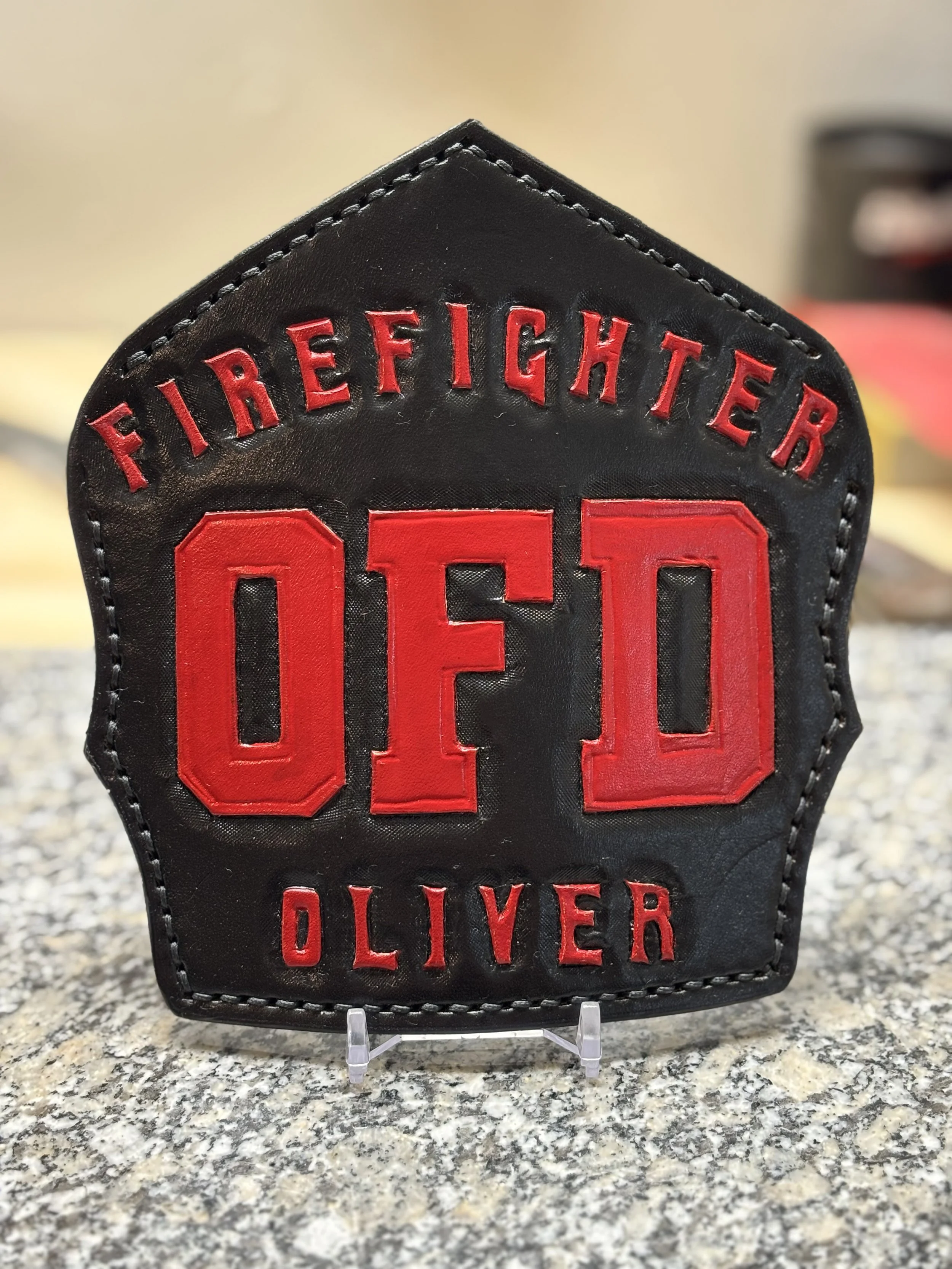 A firefighter badge with the name Oliver, featuring black leather with red embroidery reading 'FIRE FIGHTER' at the top, 'OFD' in the center, and 'OLIVER' at the bottom.