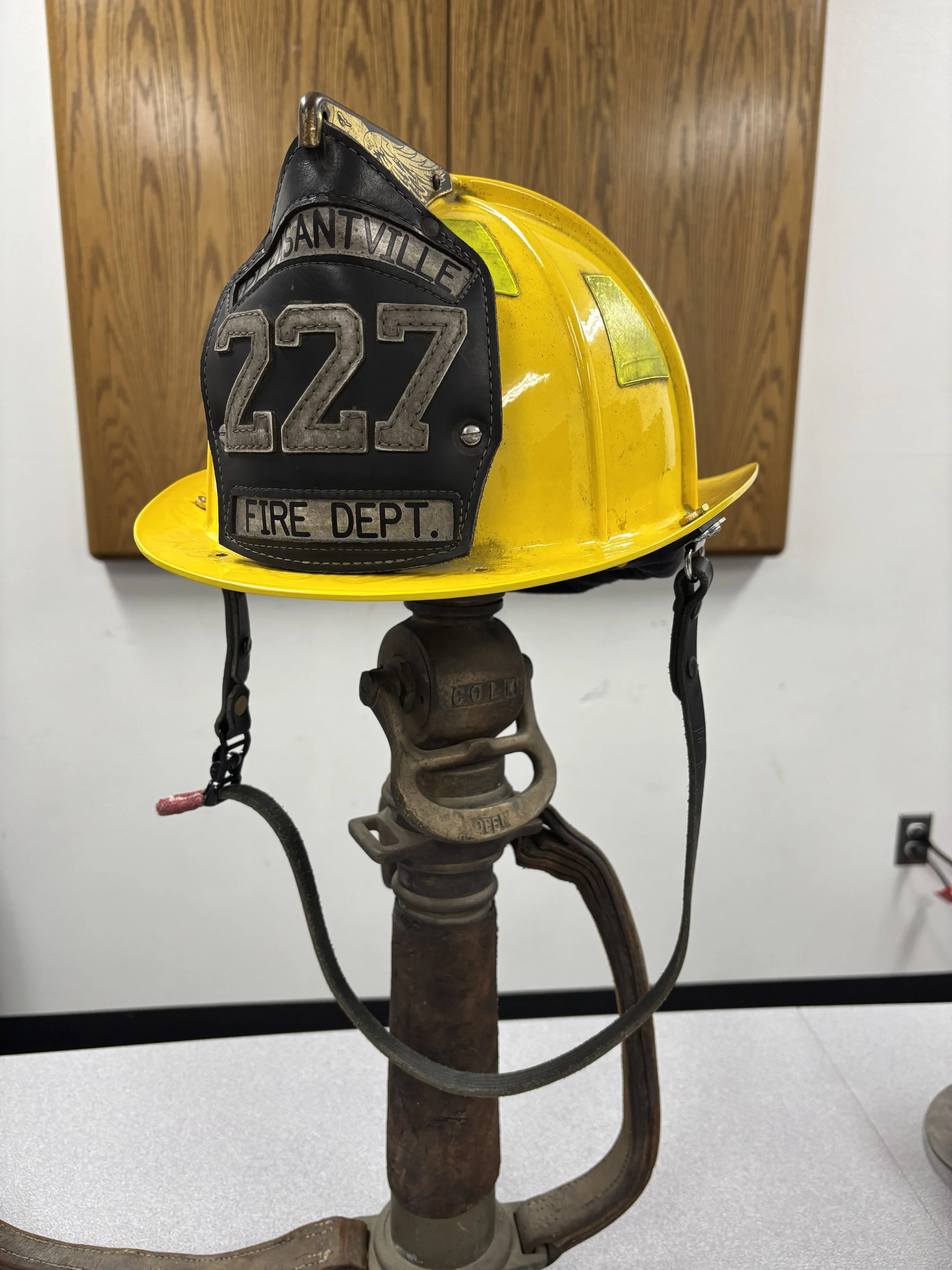 A yellow firefighter helmet with a black leather patch labeled 'SANT VILLE 227 FIRE DEPT.' is displayed on an old firefighter apparatus, with a wooden panel in the background.
