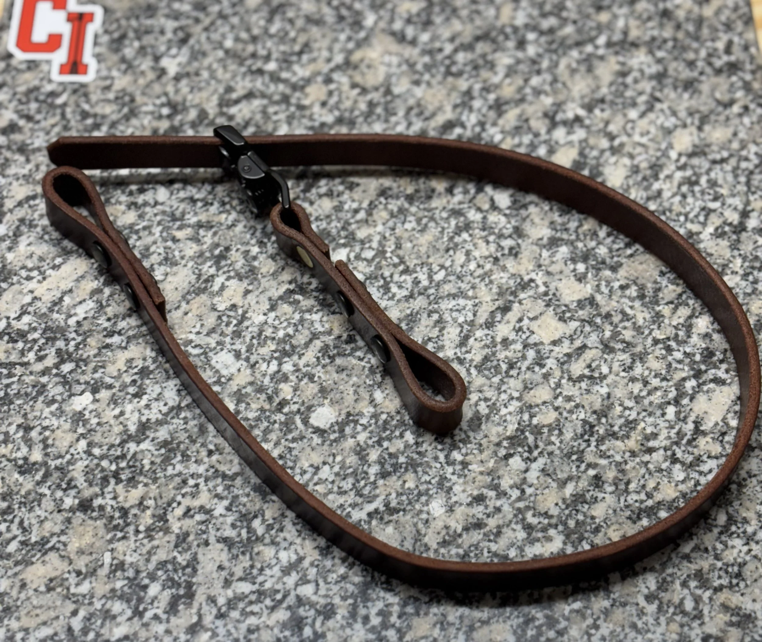 A brown leather glasses strap with black metal clips on a gray granite countertop.
