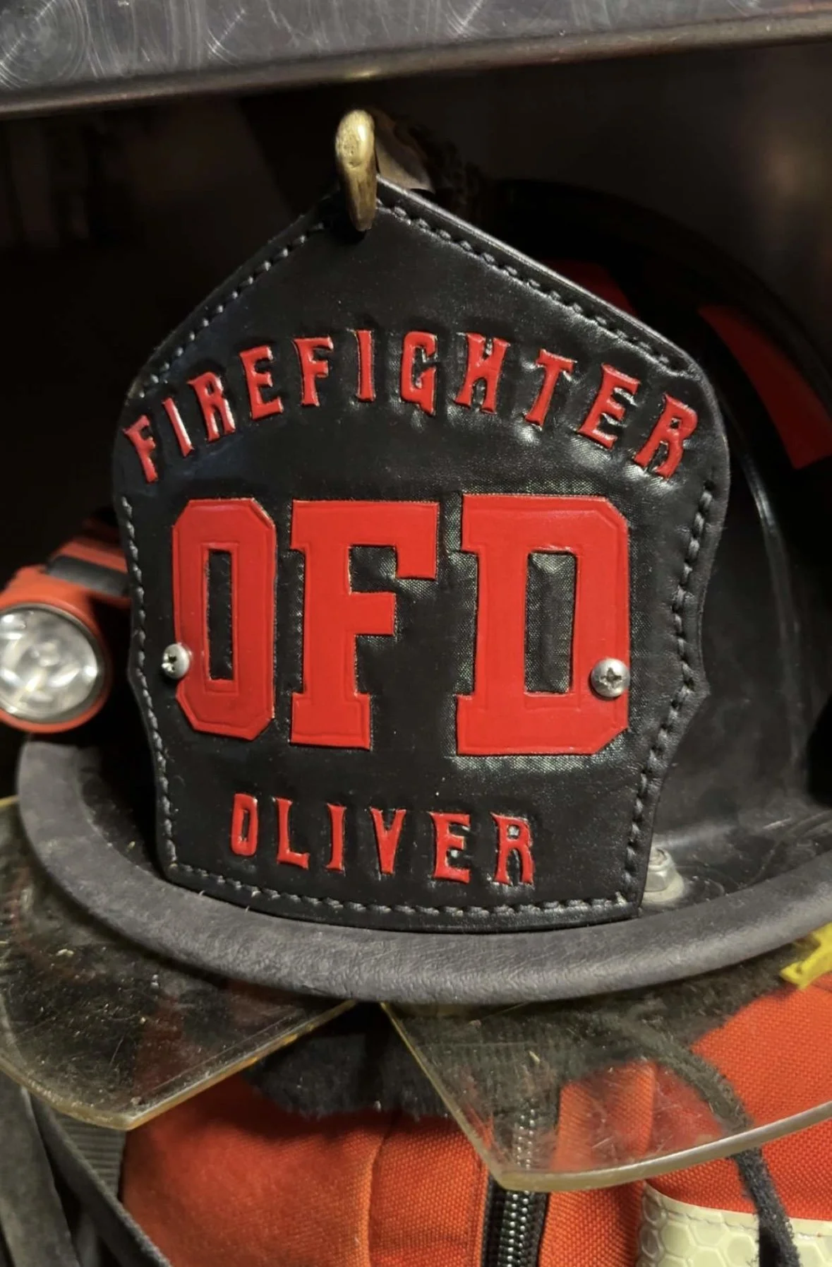 A black leather patch with red embroidered text reading 'FIRE FIGHTER O.D. OLIVER' on a firefighter's gear.