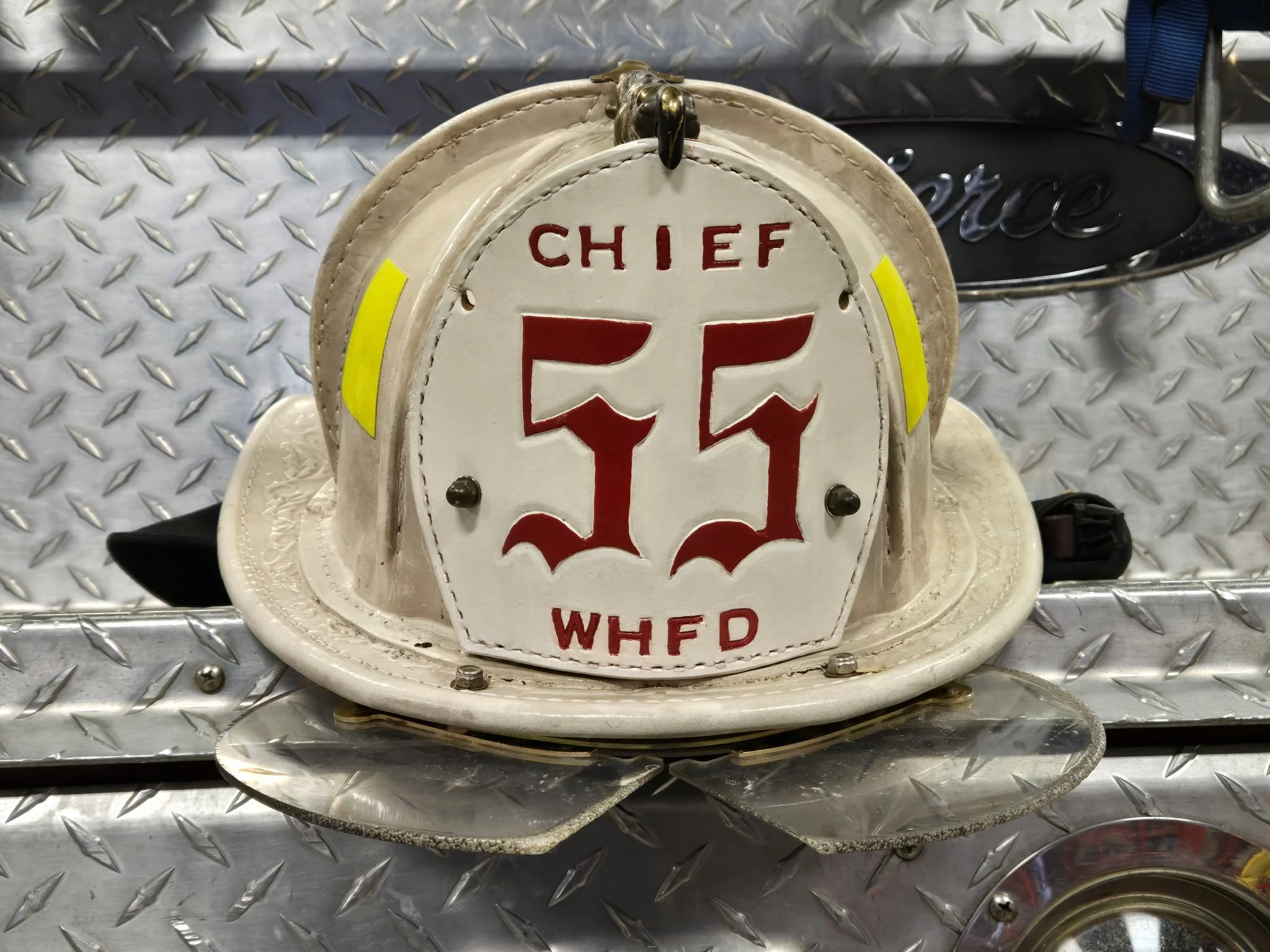 White firefighter helmet with red emblem reading 'Chief 11' and 'WHFD', placed on a diamond-plate surface with a metal sign in the background.