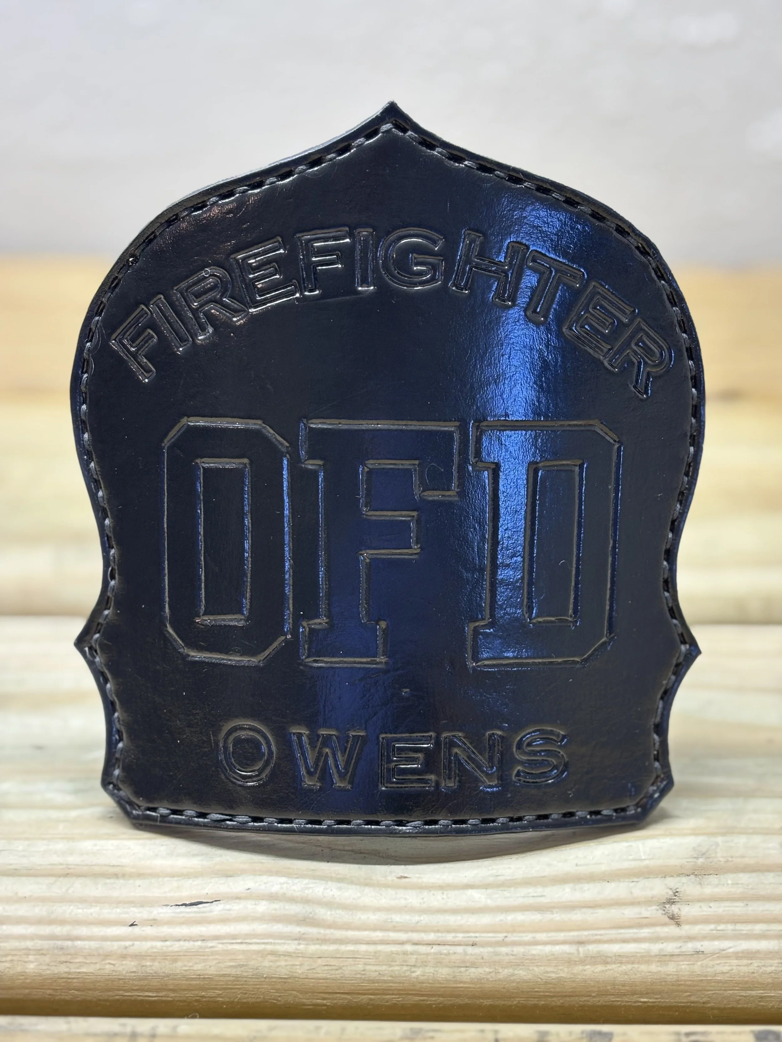 Black leather badge with embossed text reading 'FIRE FIGHTER', initials 'OFD', and the name 'OWENS' on a wooden surface.