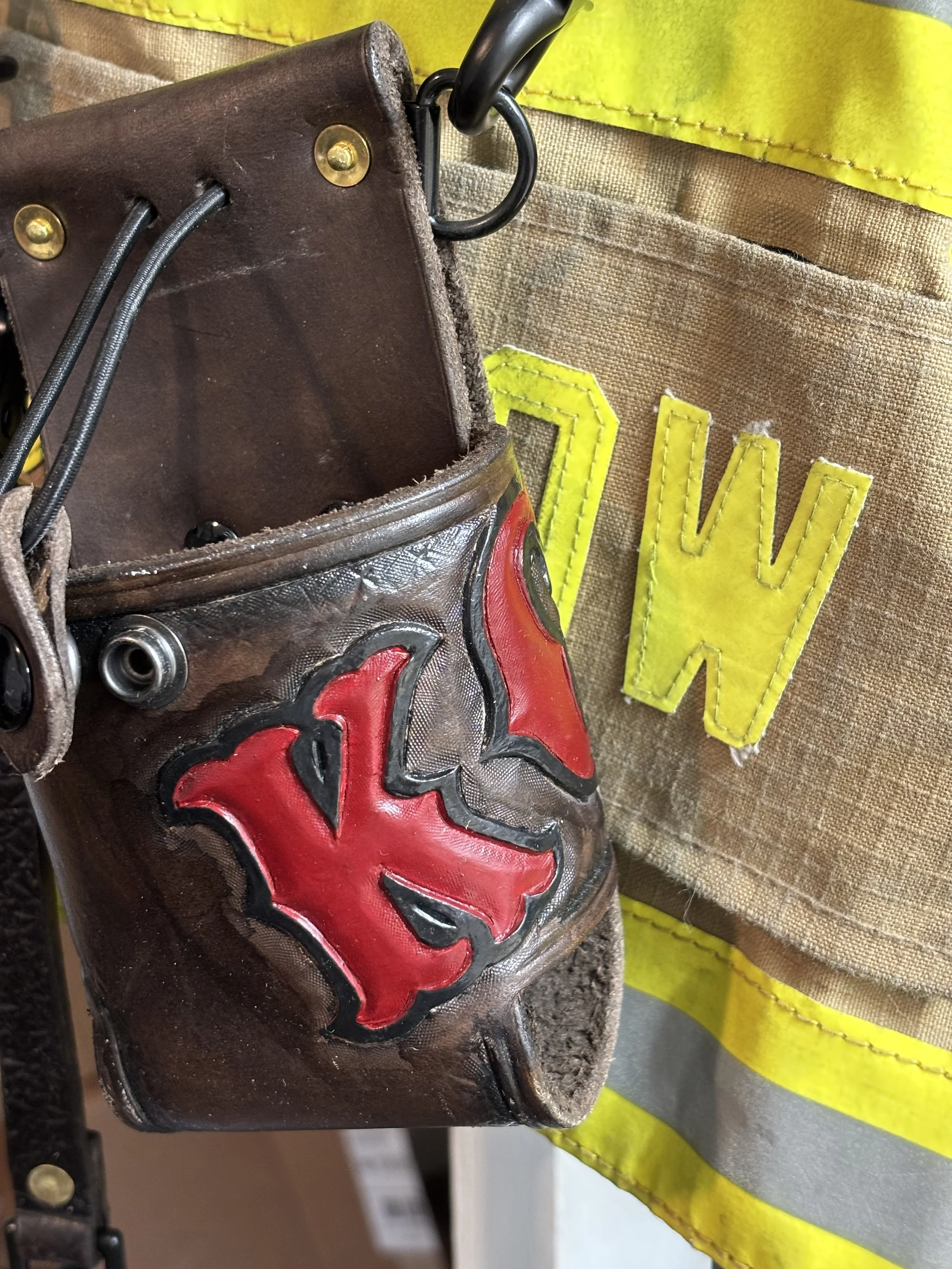 A firefighter's tool pouch with a red and black emblem of a demon face, hanging behind a yellow and reflective safety vest.