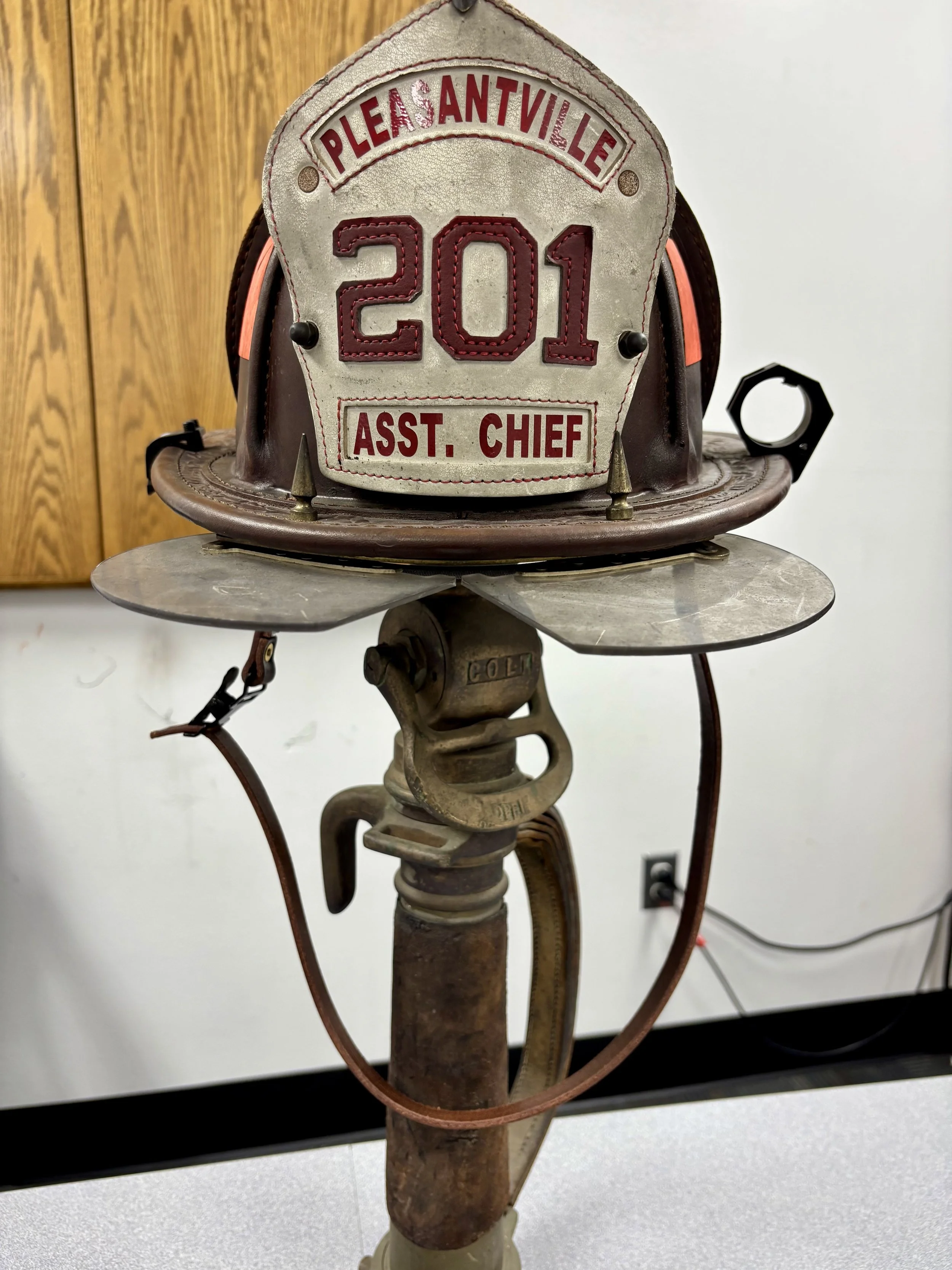 Firefighter's helmet with a firefighter's badge on top, mounted on an old fireman's axe, with a wood-paneled wall in the background.