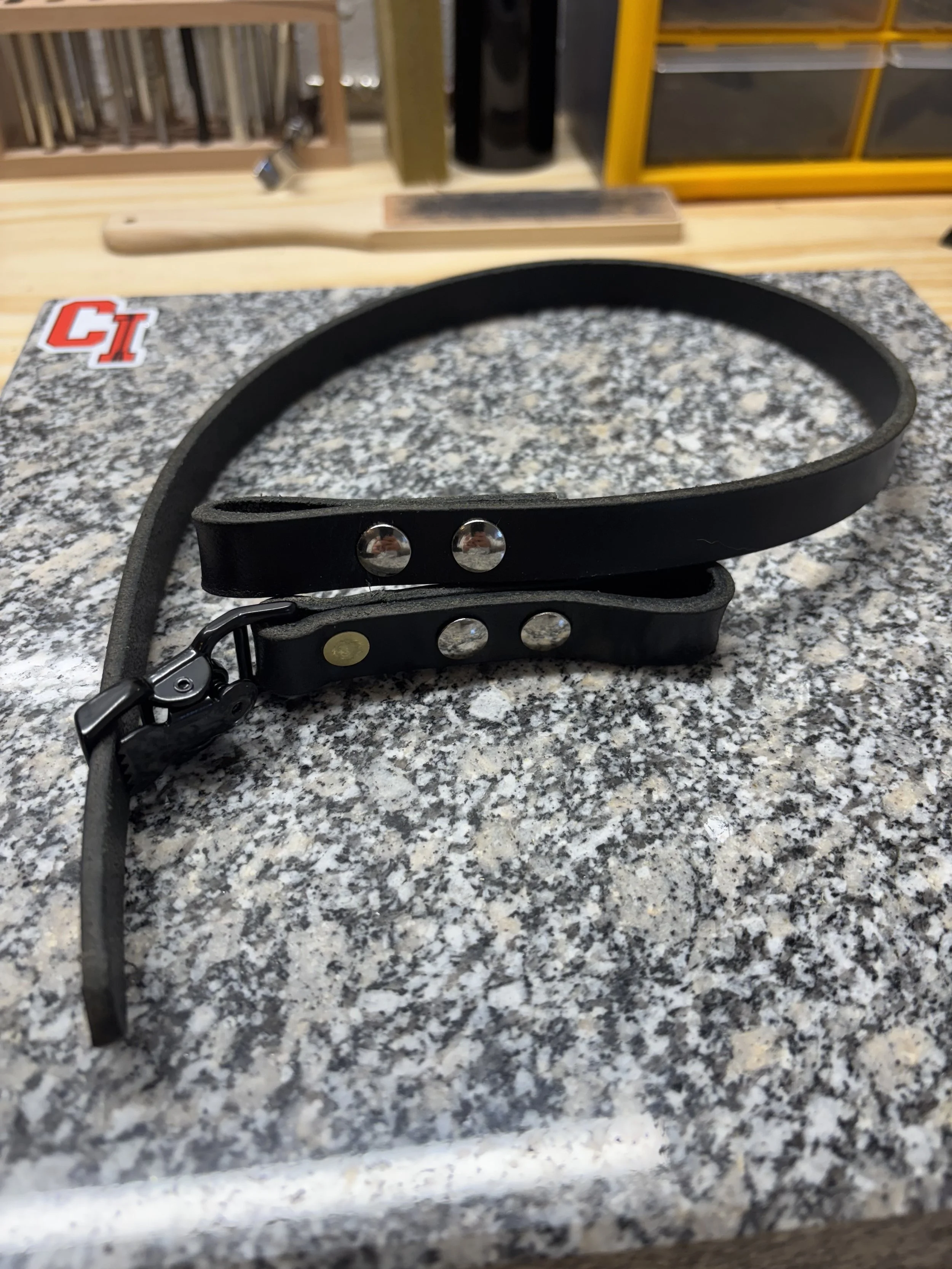A black leather dog collar with silver studs on a speckled gray work surface, with woodworking tools in the background.
