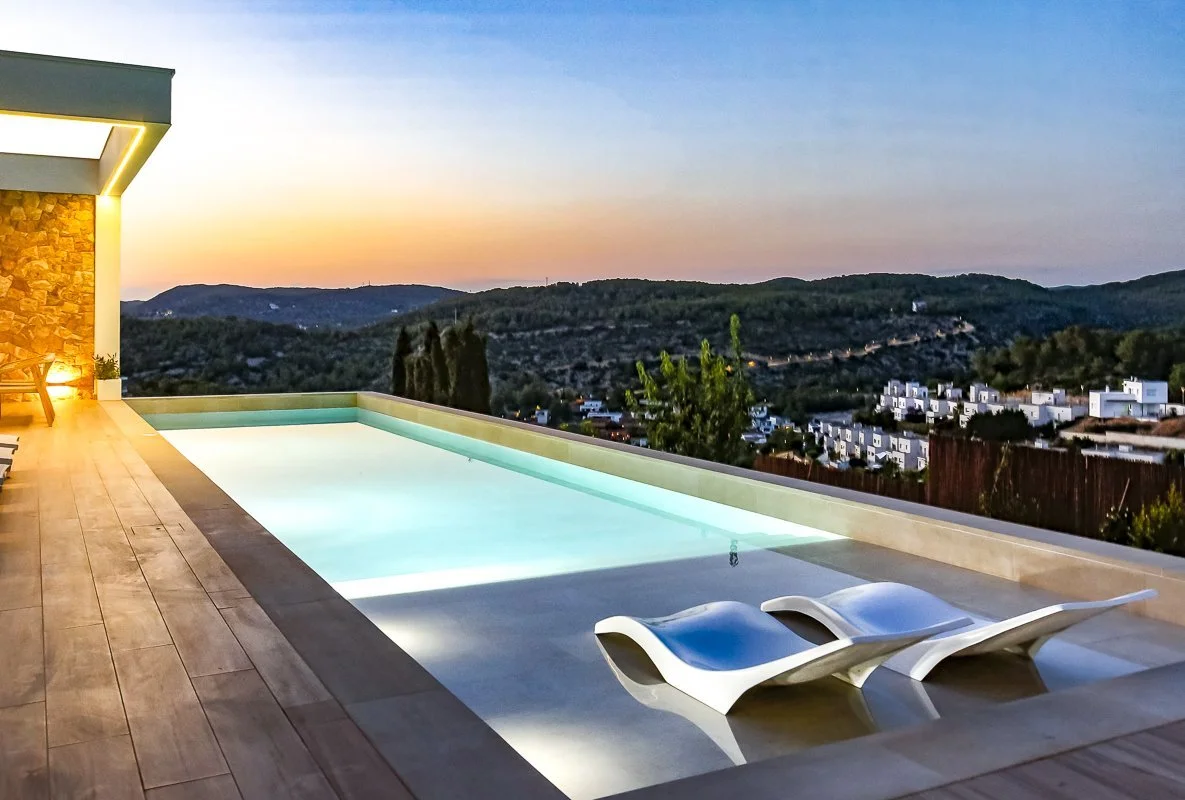 Modern rooftop infinity pool with two white lounge chairs overlooking a valley with rolling hills and houses at sunset.