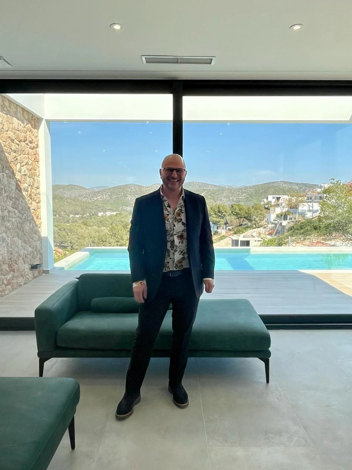 A man in a suit with a floral shirt smiling indoors in front of a large window with a view of a swimming pool, trees, and hills.