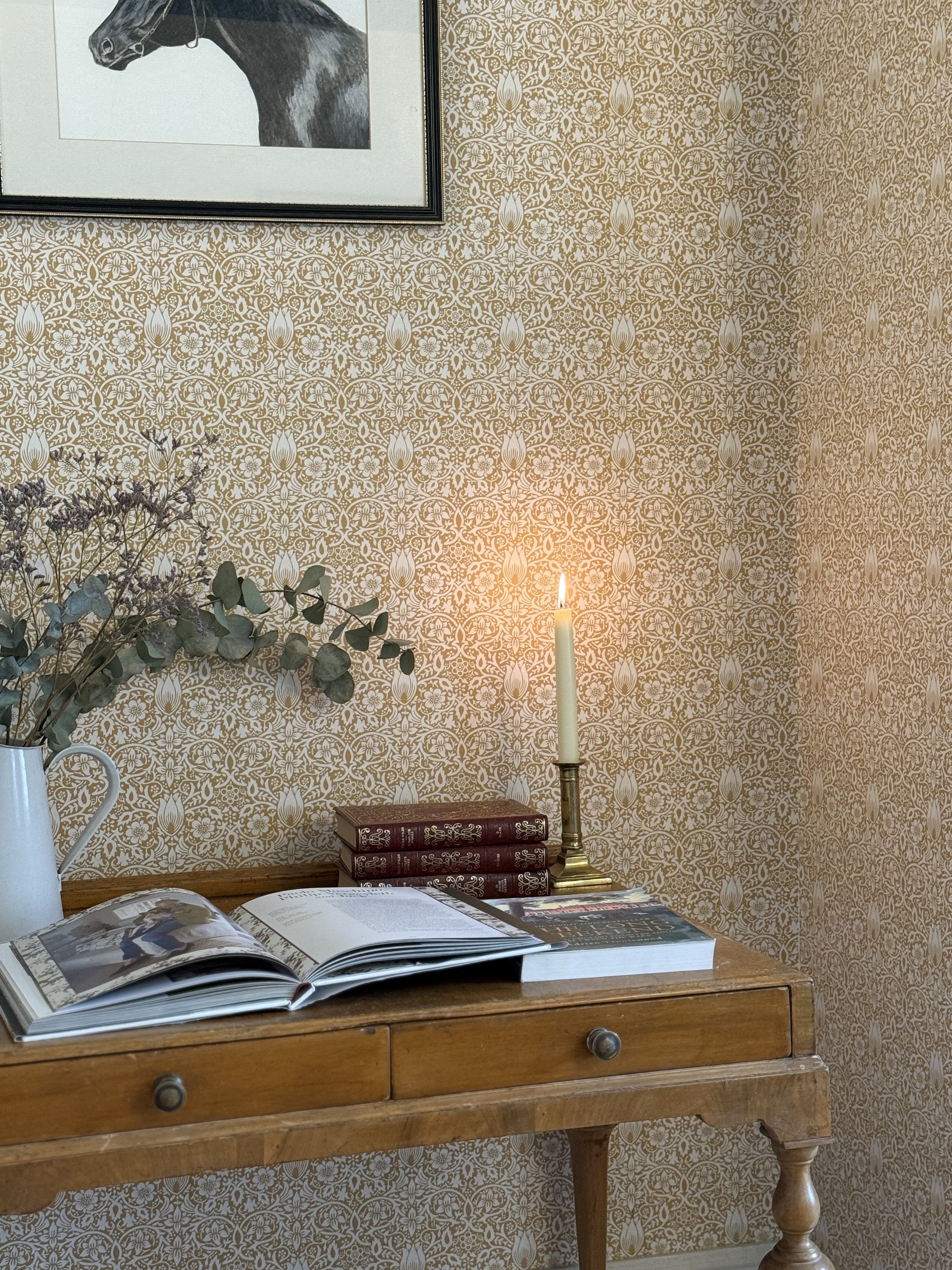 A wooden desk with an open book, a stack of three closed books, and a lit candle in a brass holder. A vase with dried flowers and eucalyptus leaves is on the left side of the desk. The background features a wallpapered wall with intricate gold and wh