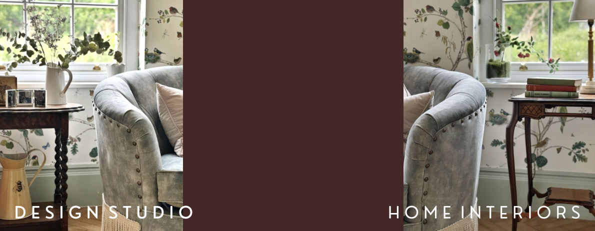 A cozy living room corner with a gray velvet armchair, floral wallpaper, window with plants, and a wooden side table with books and a lamp.