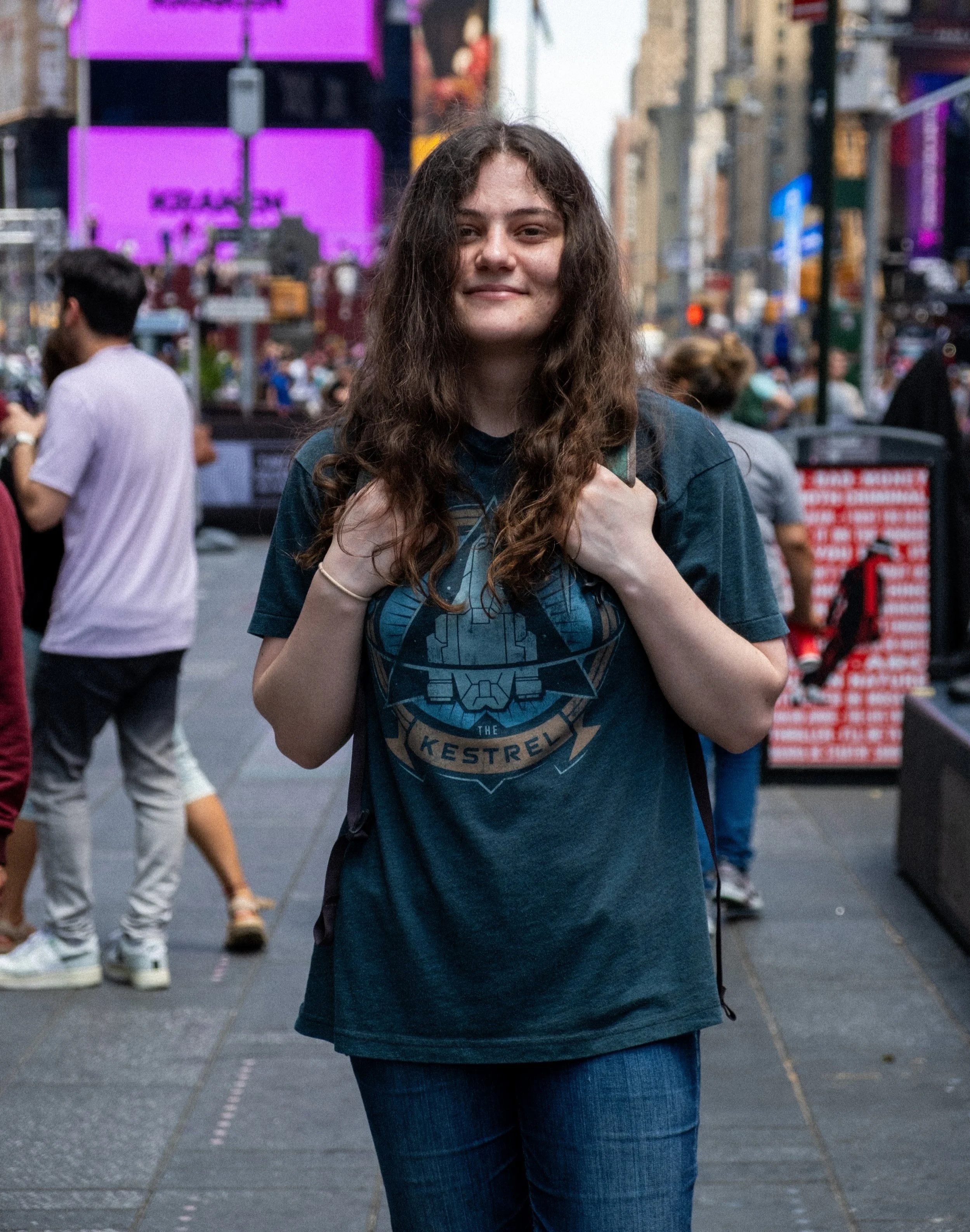 A young woman with long, curly brown hair standing on a busy city street, wearing a dark graphic T-shirt and blue jeans, with a slight smile, holding the strap of her backpack.