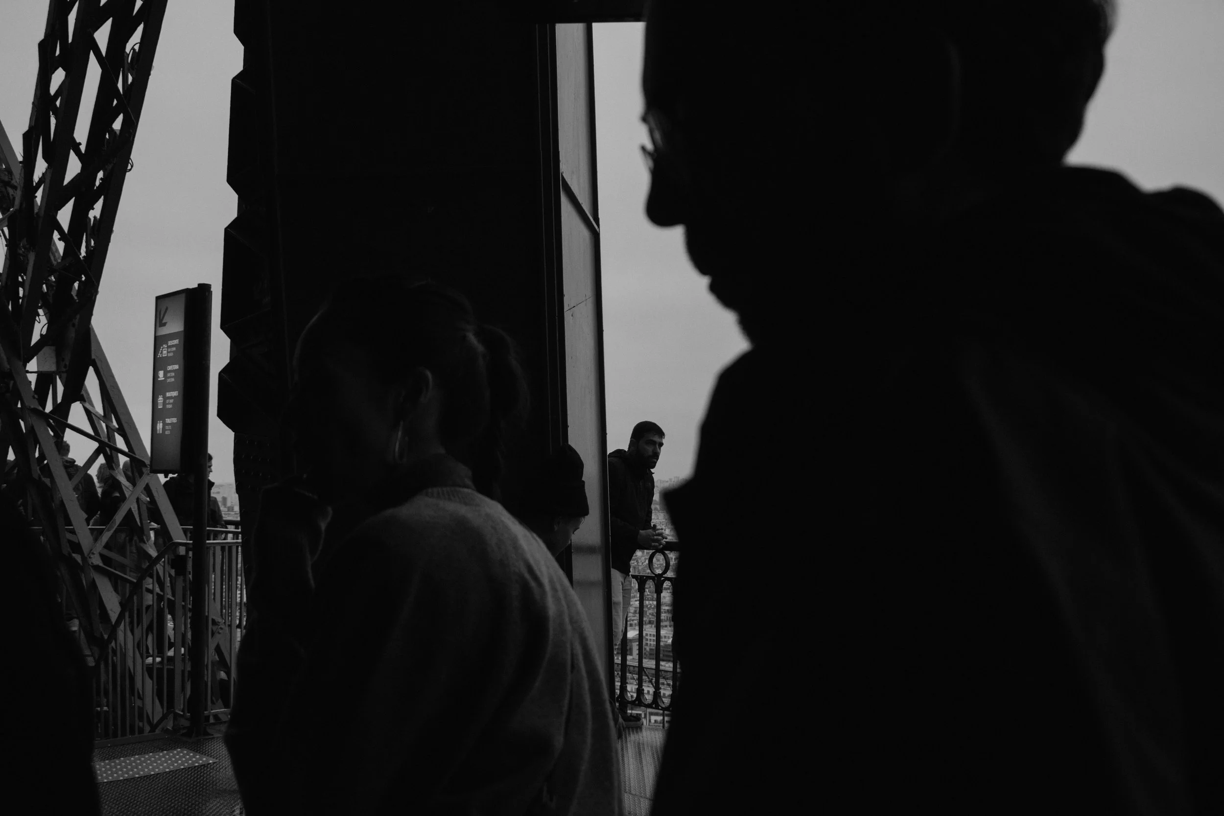 People standing on a balcony overlooking a city, with some silhouettes visible and a man in the background leaning on the railing, in black and white.