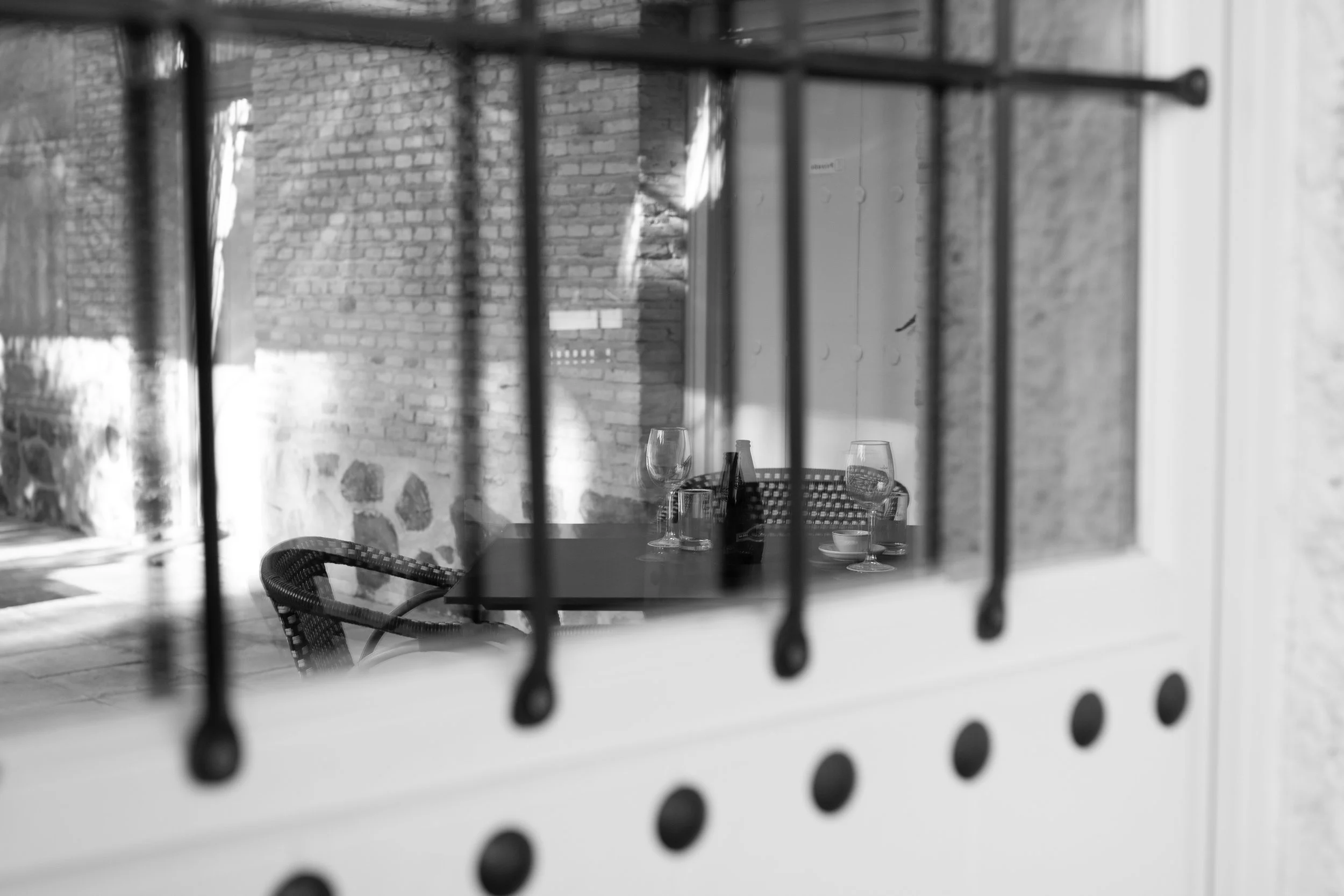A black and white photo of a table with glasses and a bottle, seen through a window with bars. The background shows a brick wall.