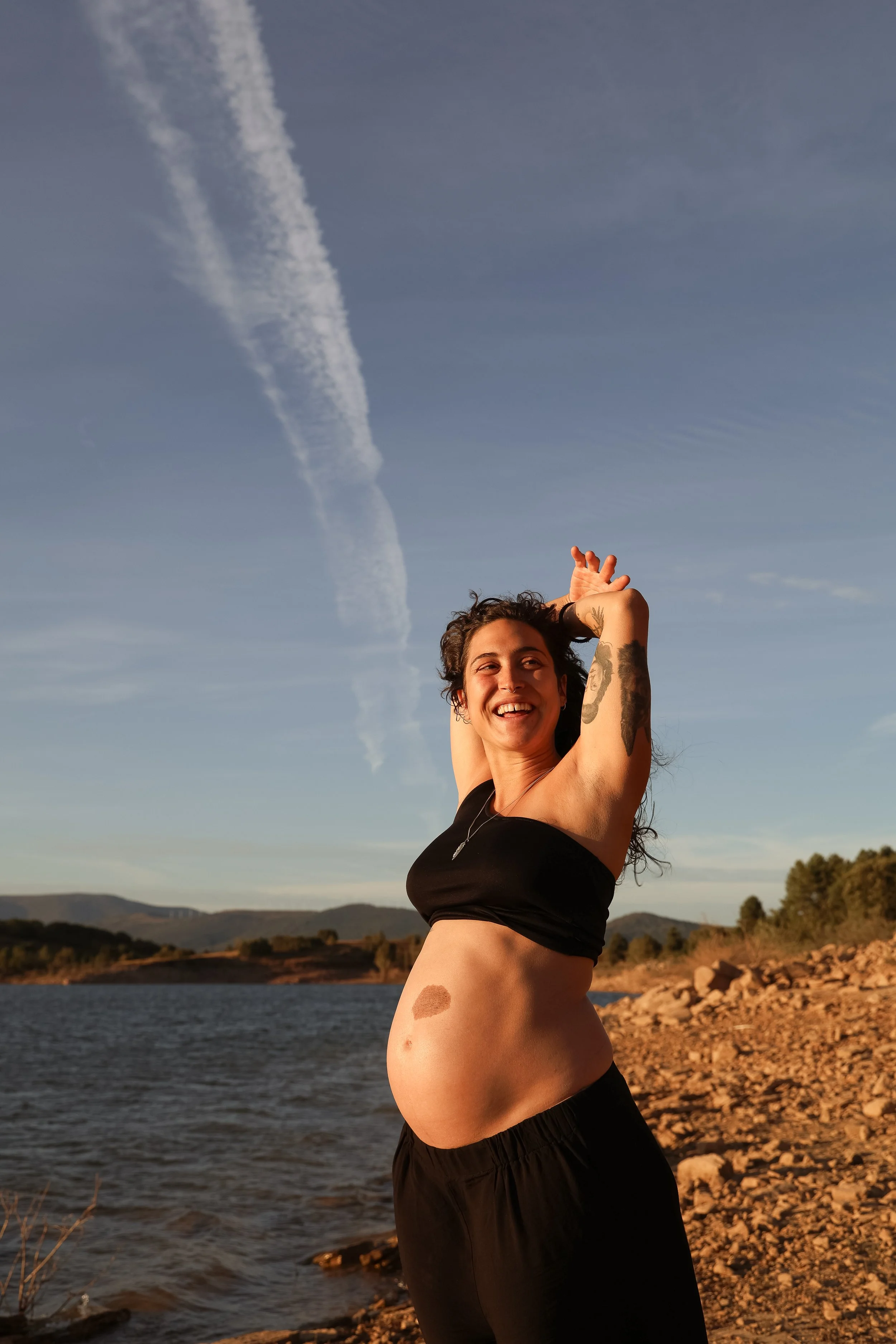A pregnant woman with tattoos standing by a body of water on a rocky shoreline, smiling with arms raised above her head, against a backdrop of blue sky and distant mountains.