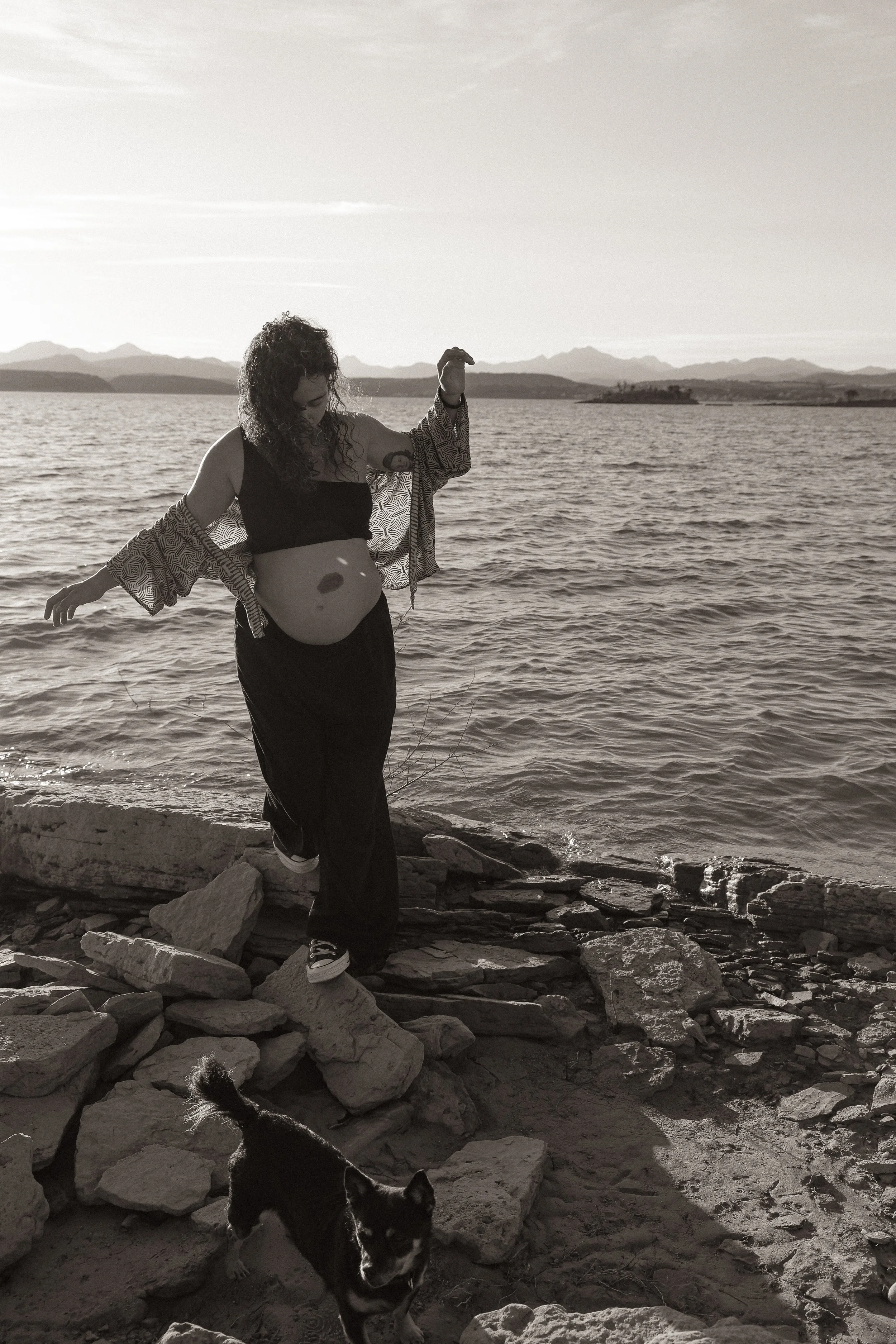 A woman with curly hair standing on a rocky shoreline next to a dog with the ocean and mountains in the background, during sunset.