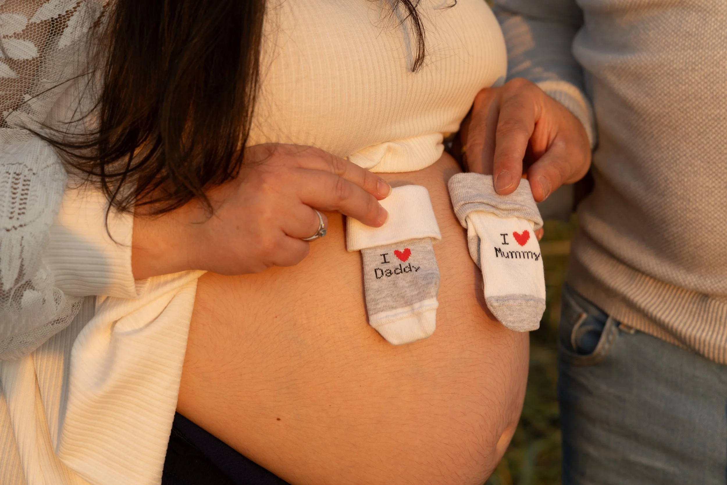 Close-up of pregnant woman's belly with two small baby socks hanging on her belly, one sock says 'I ❤️ Daddy' and the other says 'I ❤️ Mummy,' with a woman and man holding the socks.