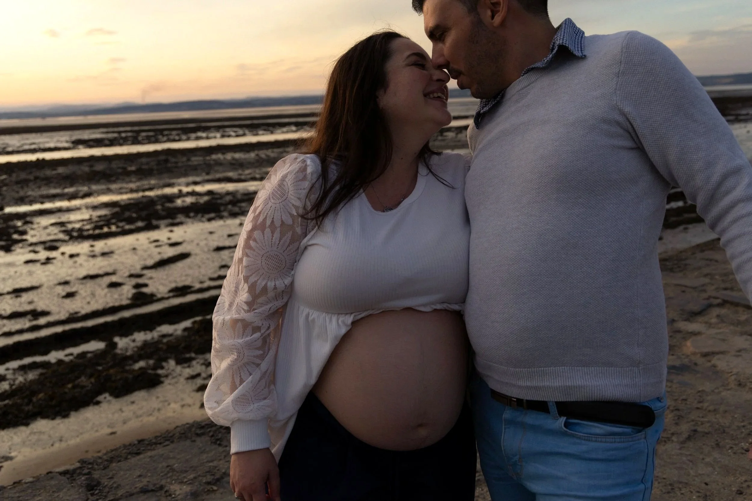 A pregnant woman and a man are standing close together on a beach at sunset, touching foreheads and smiling.