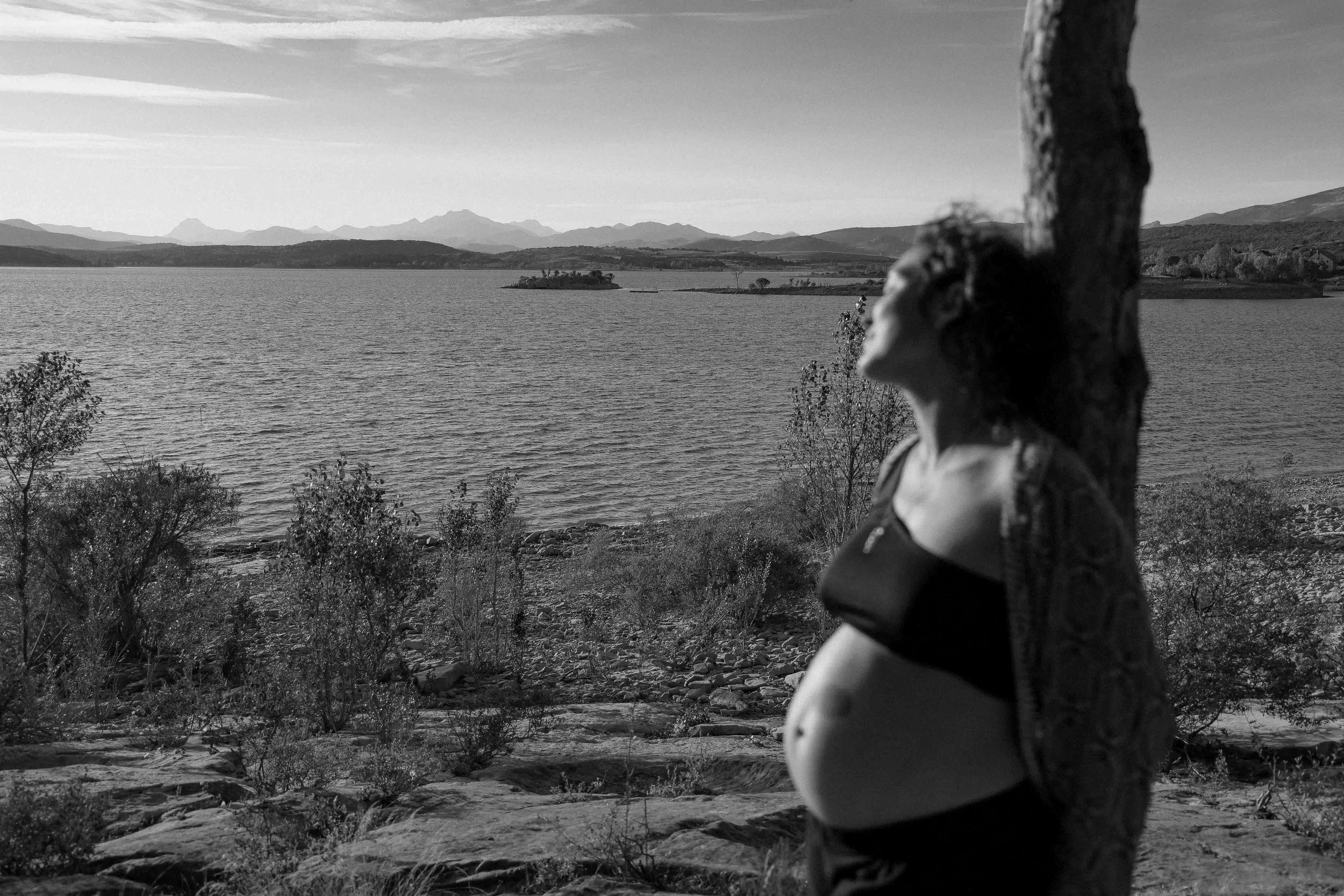 fine art image of a pregnant woman with curly hair wearing a black crop top and black pants walking along a rocky shoreline by a body of water, with hills in the background, in black and white.