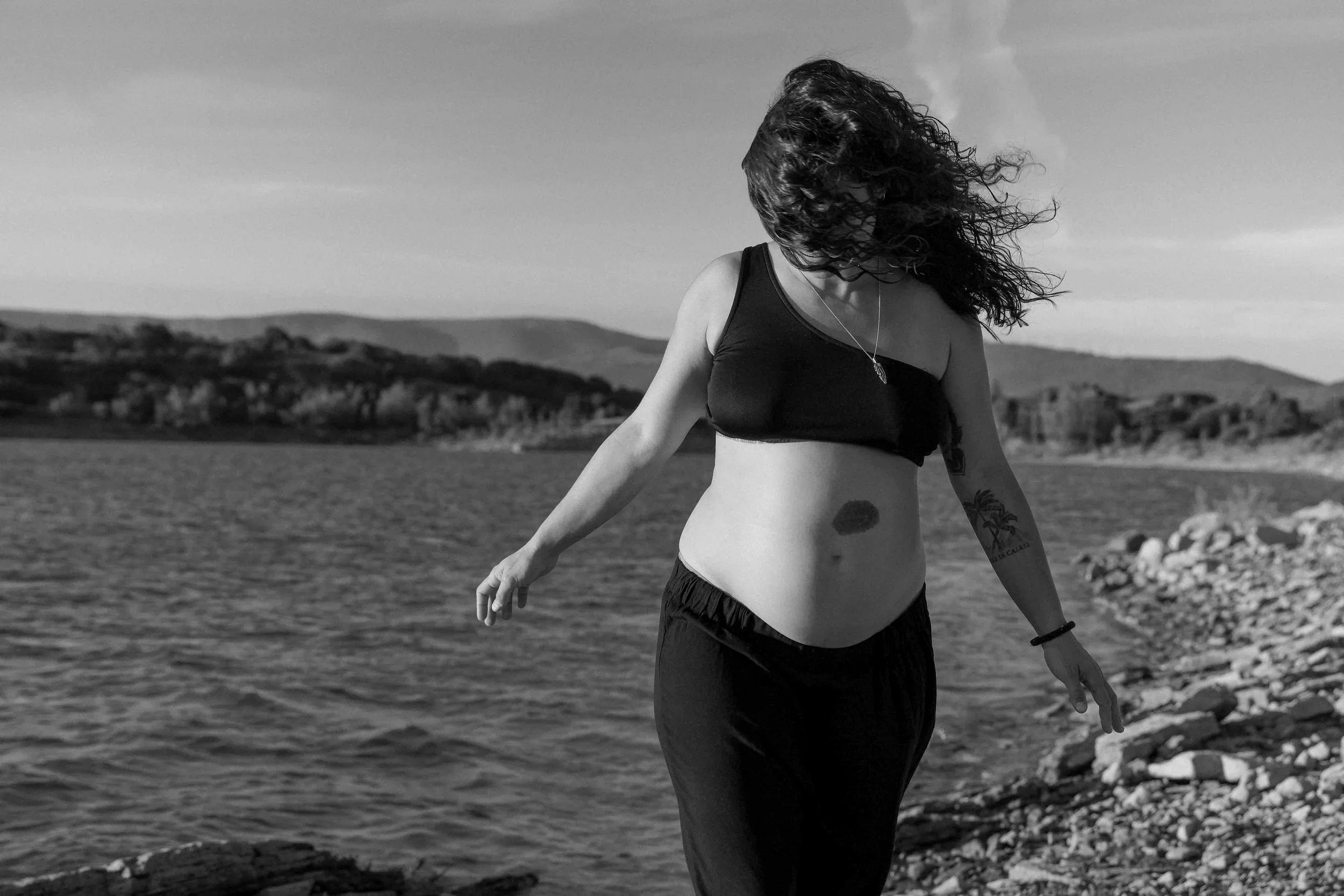 fine art image of a pregnant woman with curly hair wearing a black crop top and black pants walking along a rocky shoreline by a body of water, with hills in the background, in black and white.