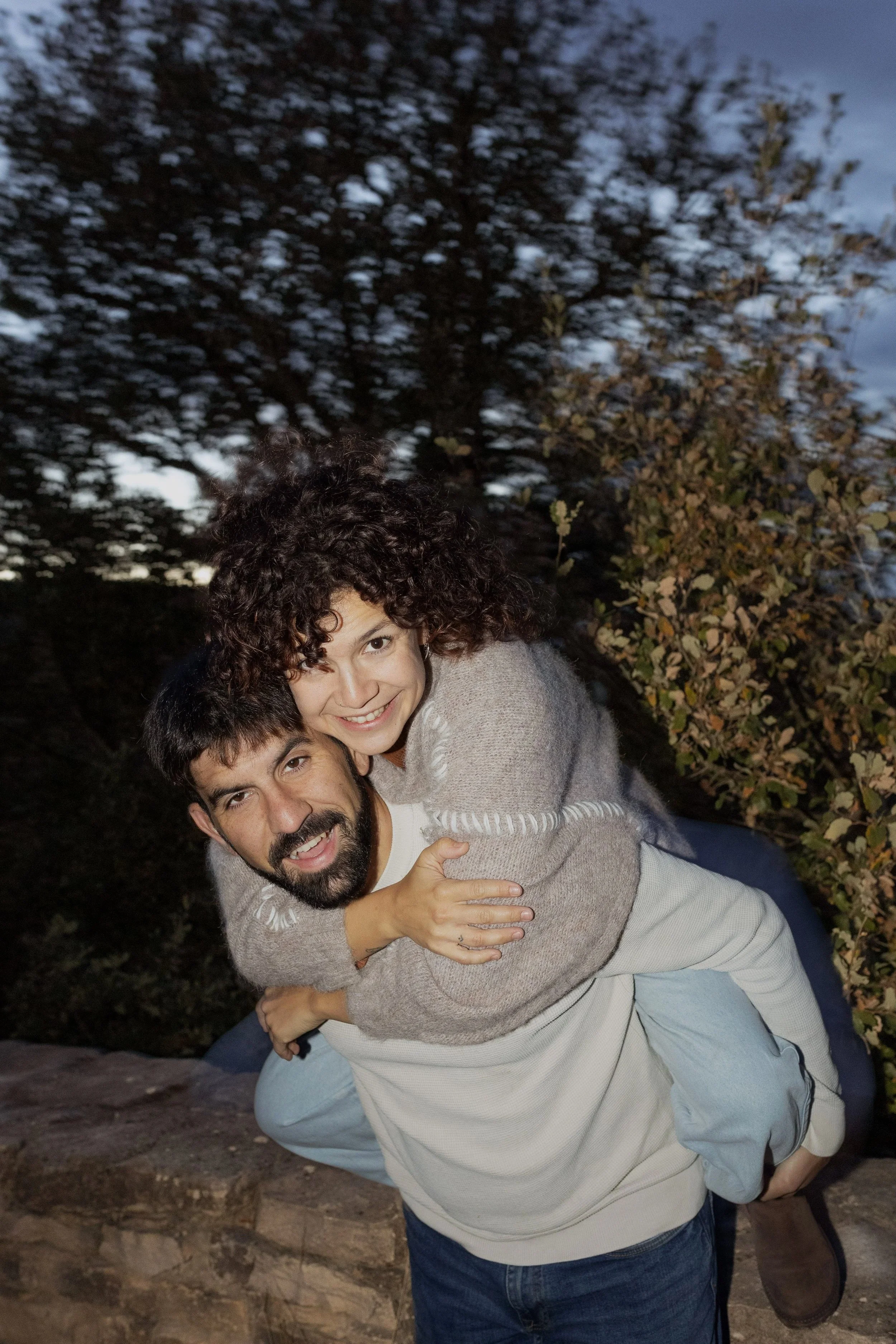 A man giving a woman a piggyback ride outside during the evening, with trees and a cloudy sky in the background.