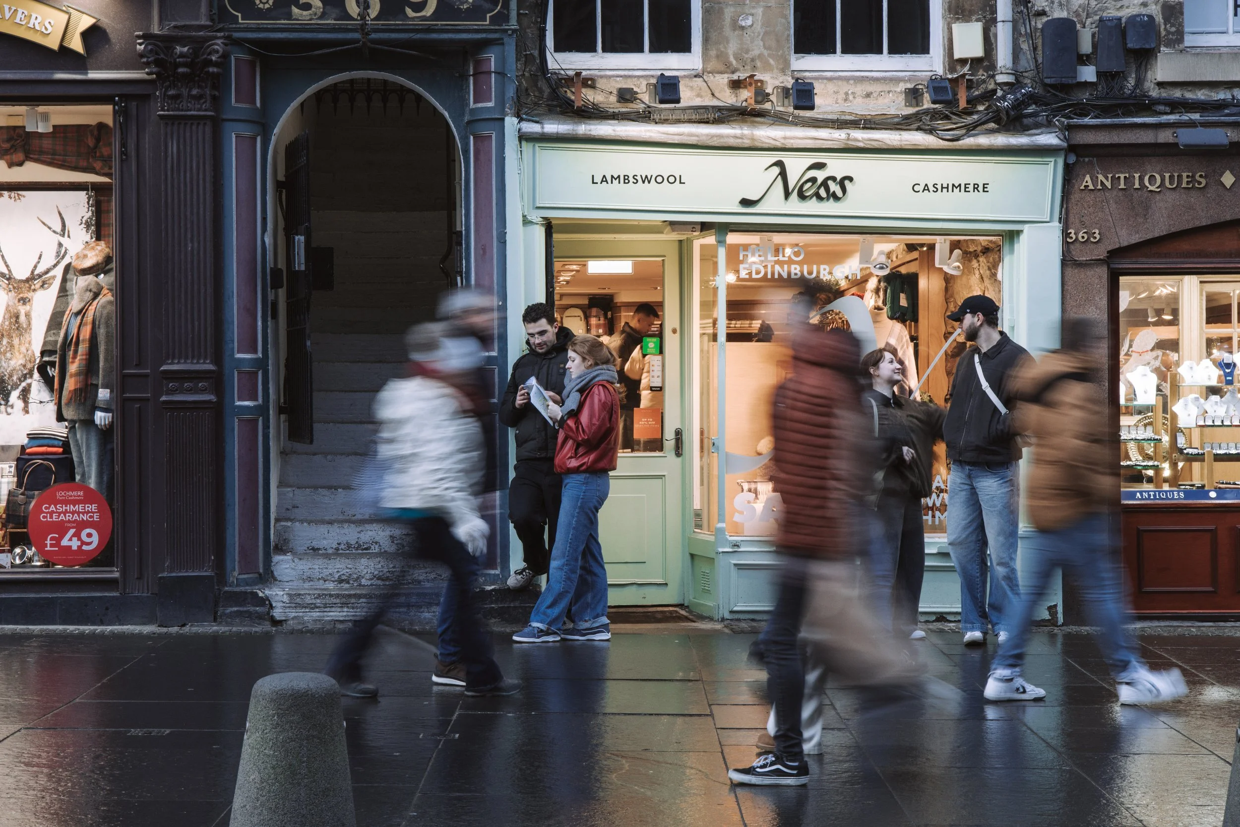 People walking on a wet city sidewalk in front of a store with a sign that says 'Ness' offering lambswool, cashmere, and antiques. Some individuals are blurred due to motion, and two people are standing and chatting near the entrance.