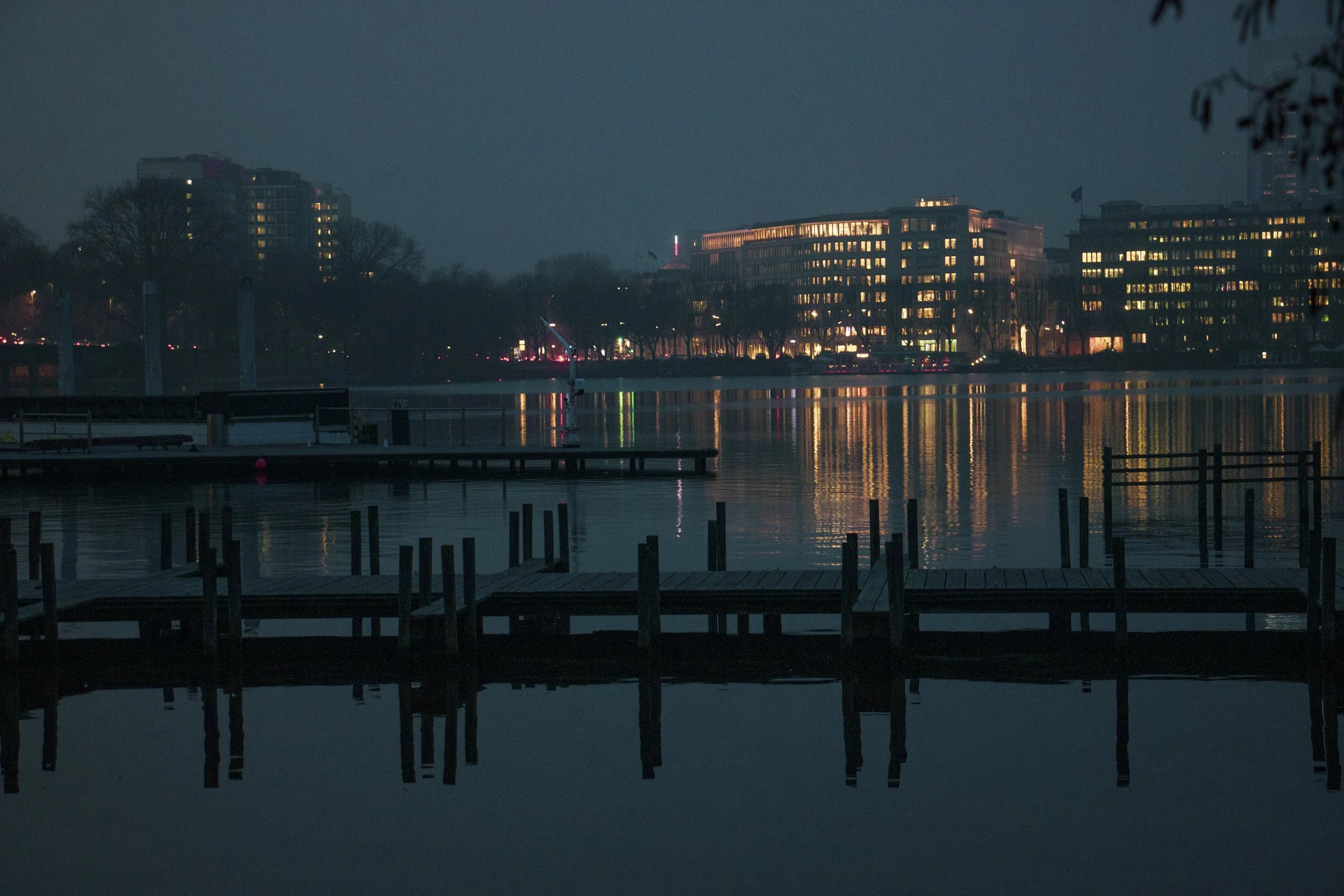 Night view of a cityscape across a body of water with illuminated buildings and reflections on the water's surface, wooden docks in the foreground.