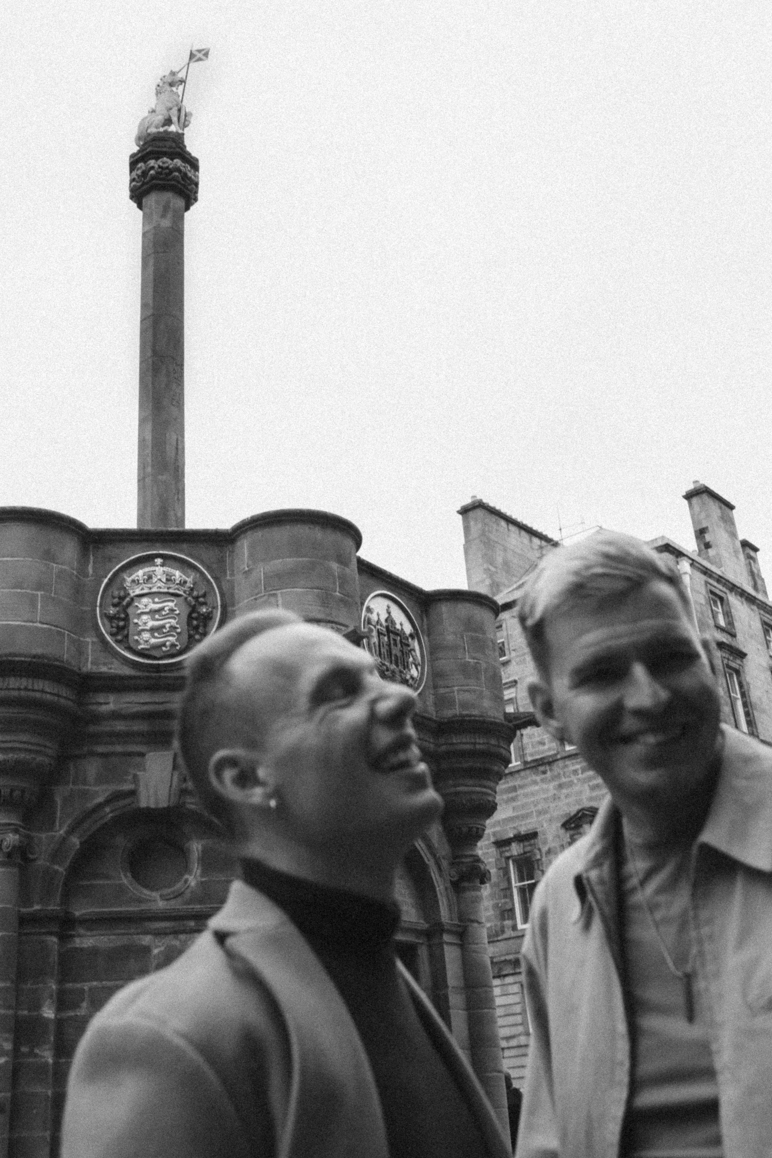 Two young men smiling and looking at each other outside historic European architecture with stone buildings and a tall column topped with an unicorn, located in the Royal Mile of Edinburgh