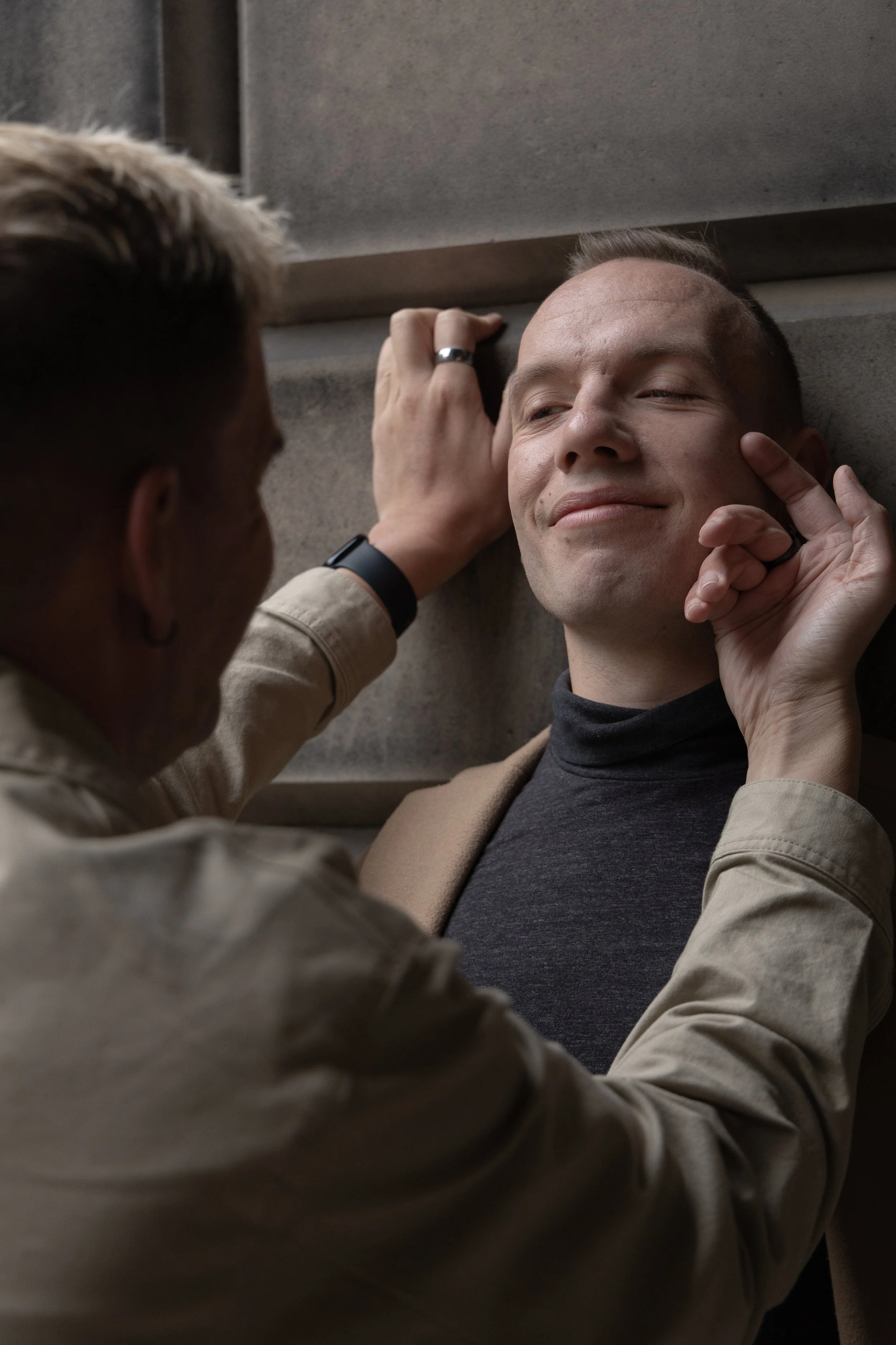 Two men on a staircase, one leaning against the wall with a smile, the other with hand on his face, showing a close-up of their interaction.